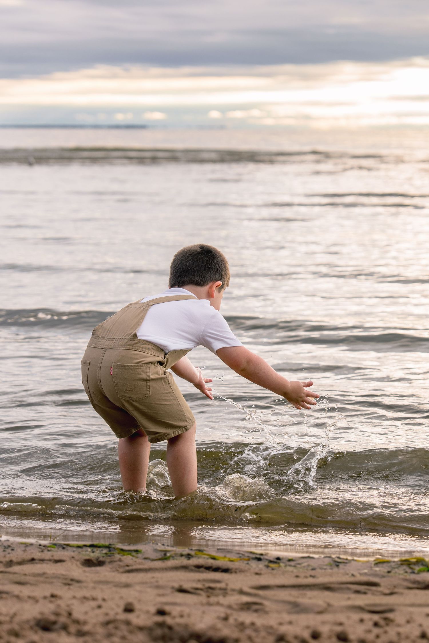 A child in khaki shorts splashes and plays in the ocean waves at sunset on a sandy beach.