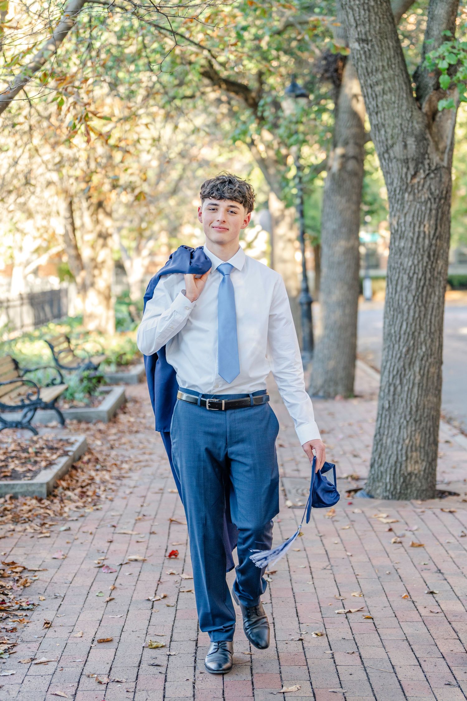 Person in white dress shirt and blue pants dancing joyfully on a brick pathway surrounded by autumn trees.