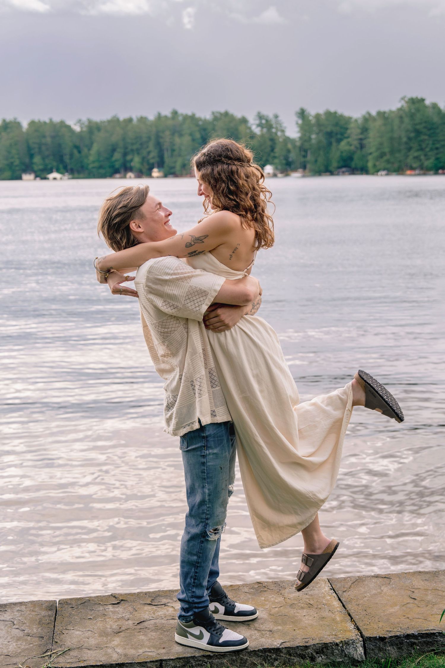 A couple stands by a lake in a romantic dance pose with trees and water in the background. Destination family photoshoot in Brantingham, NY