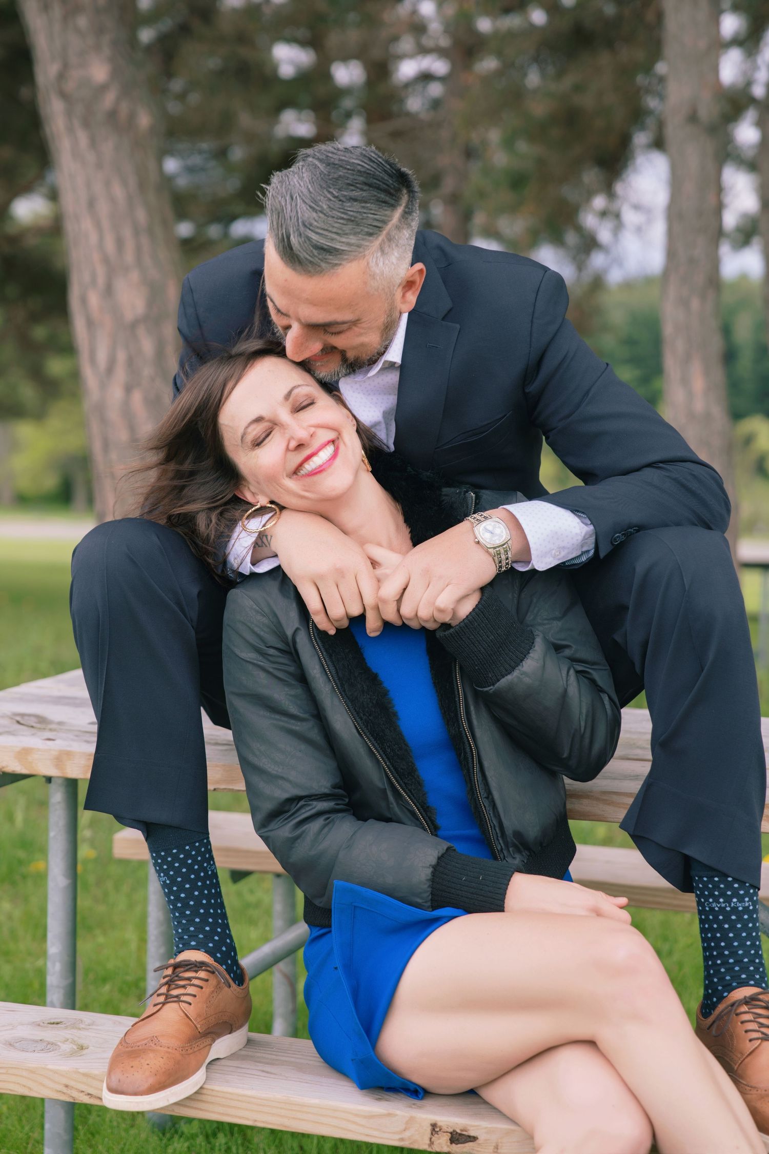 A couple in formal attire shares playful moments on a wooden bench in a park setting.