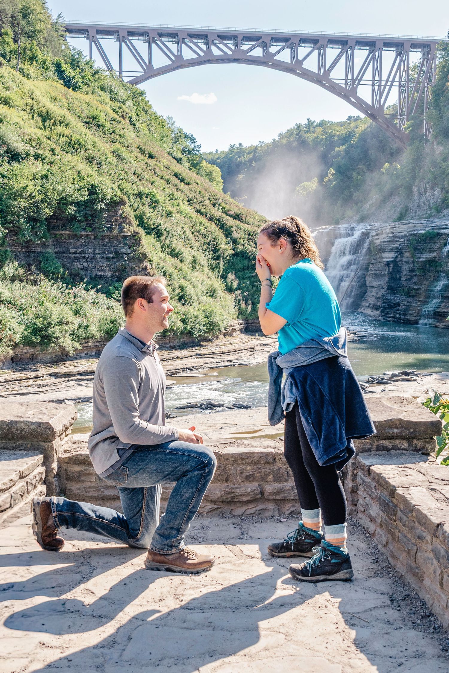Two people sit on a stone wall overlooking Letchworth State Park gorge with a railroad bridge visible in the background.