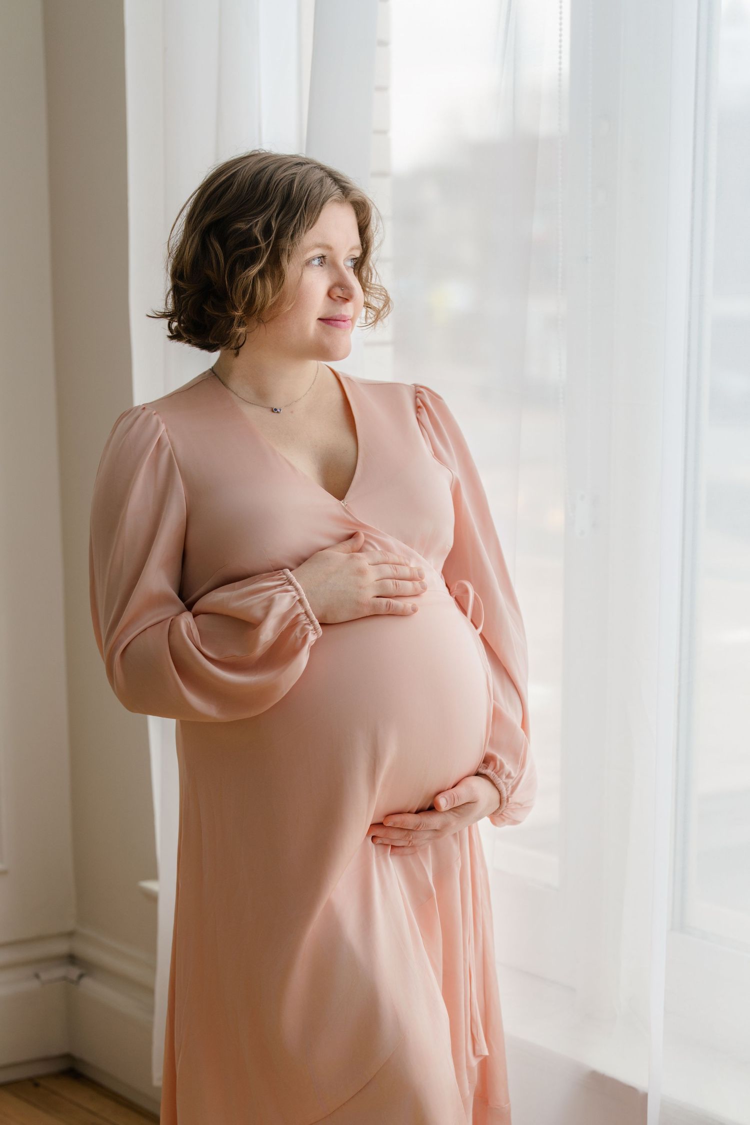 Pregnant woman in blush pink dress stands near window with natural light, gently holding her baby bump.