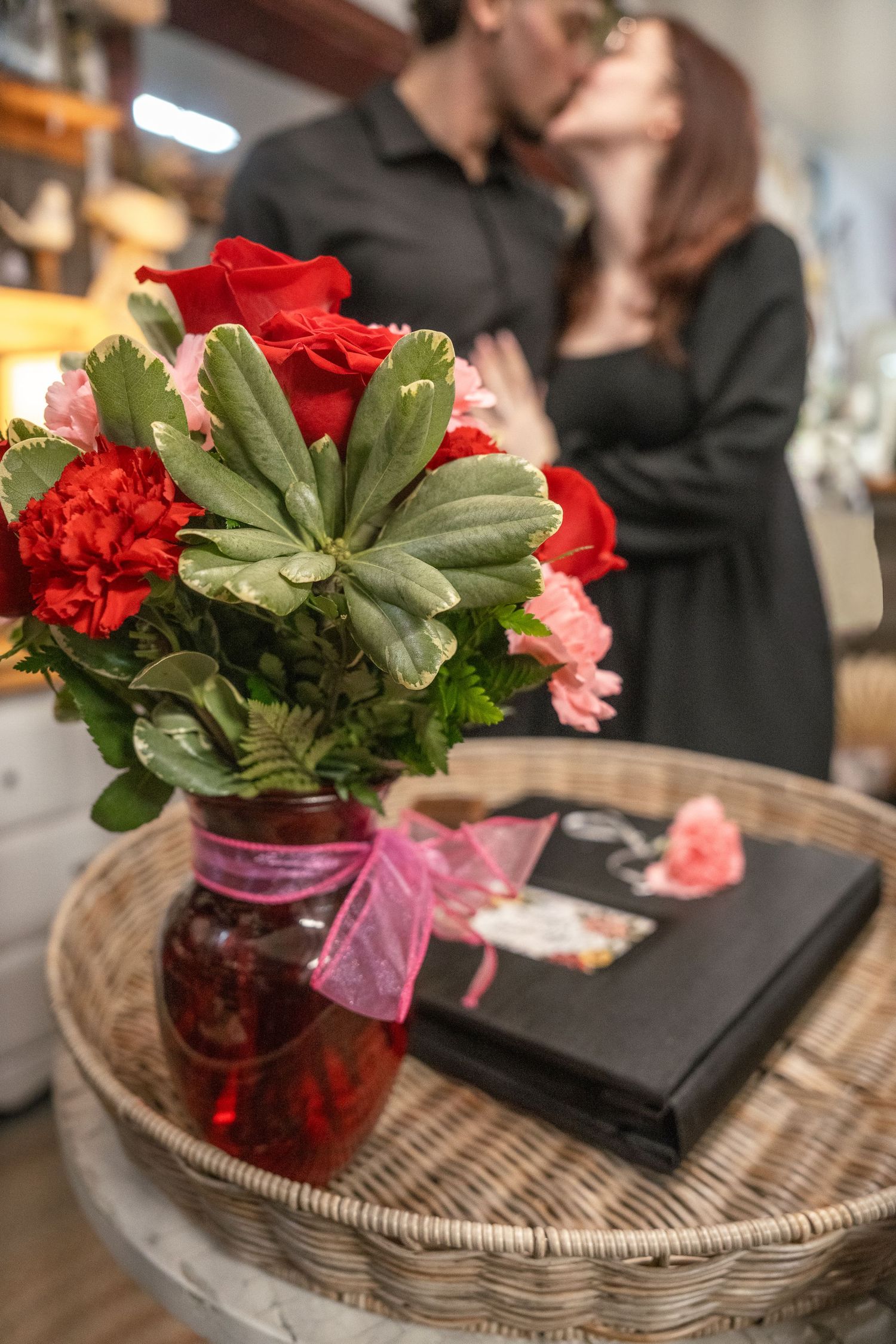 A romantic couple kissing behind a red vase bouquet of roses and carnations with a gift book on a wicker table.