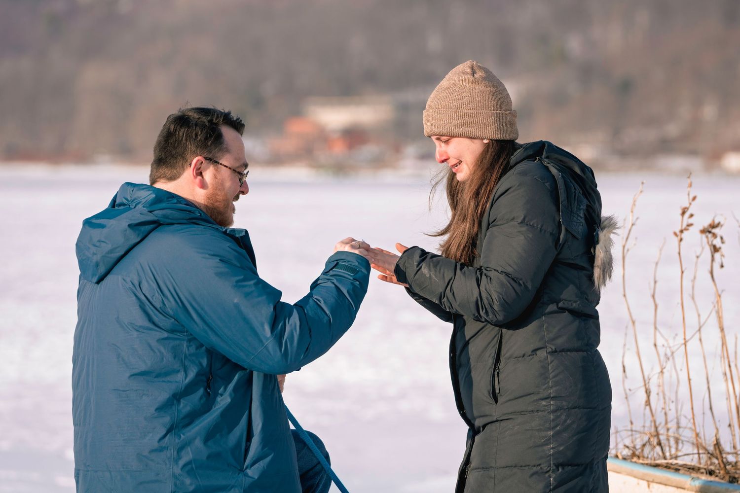 Man on one knee proposing to woman outdoors in snowy winter landscape near frozen lake.
