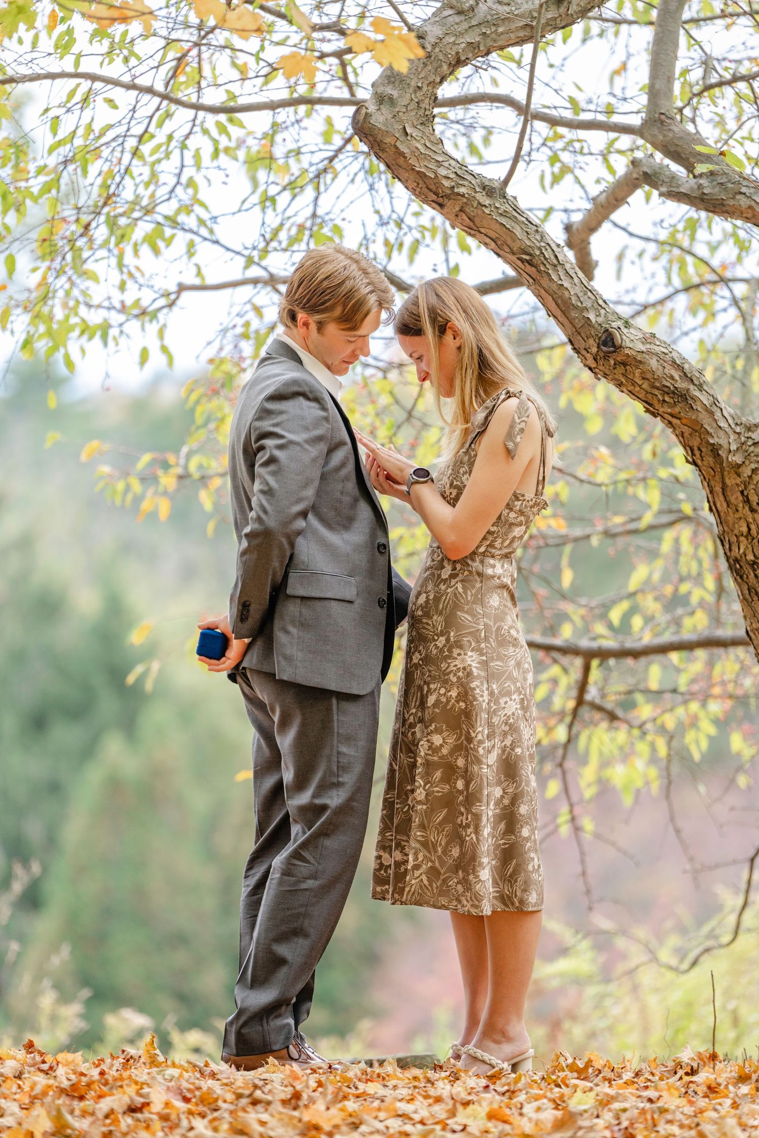 A couple stands under a tree with autumn leaves on the ground in a romantic outdoor setting.