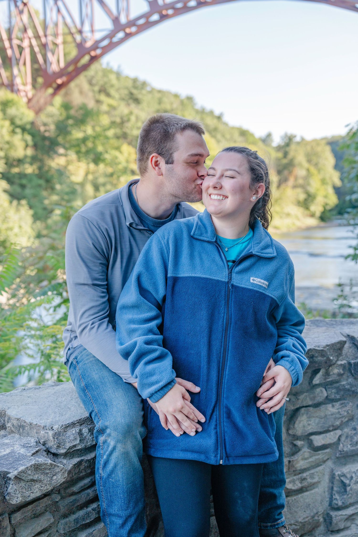 A couple shares an intimate moment near a bridge with a scenic river and trees in the background.
