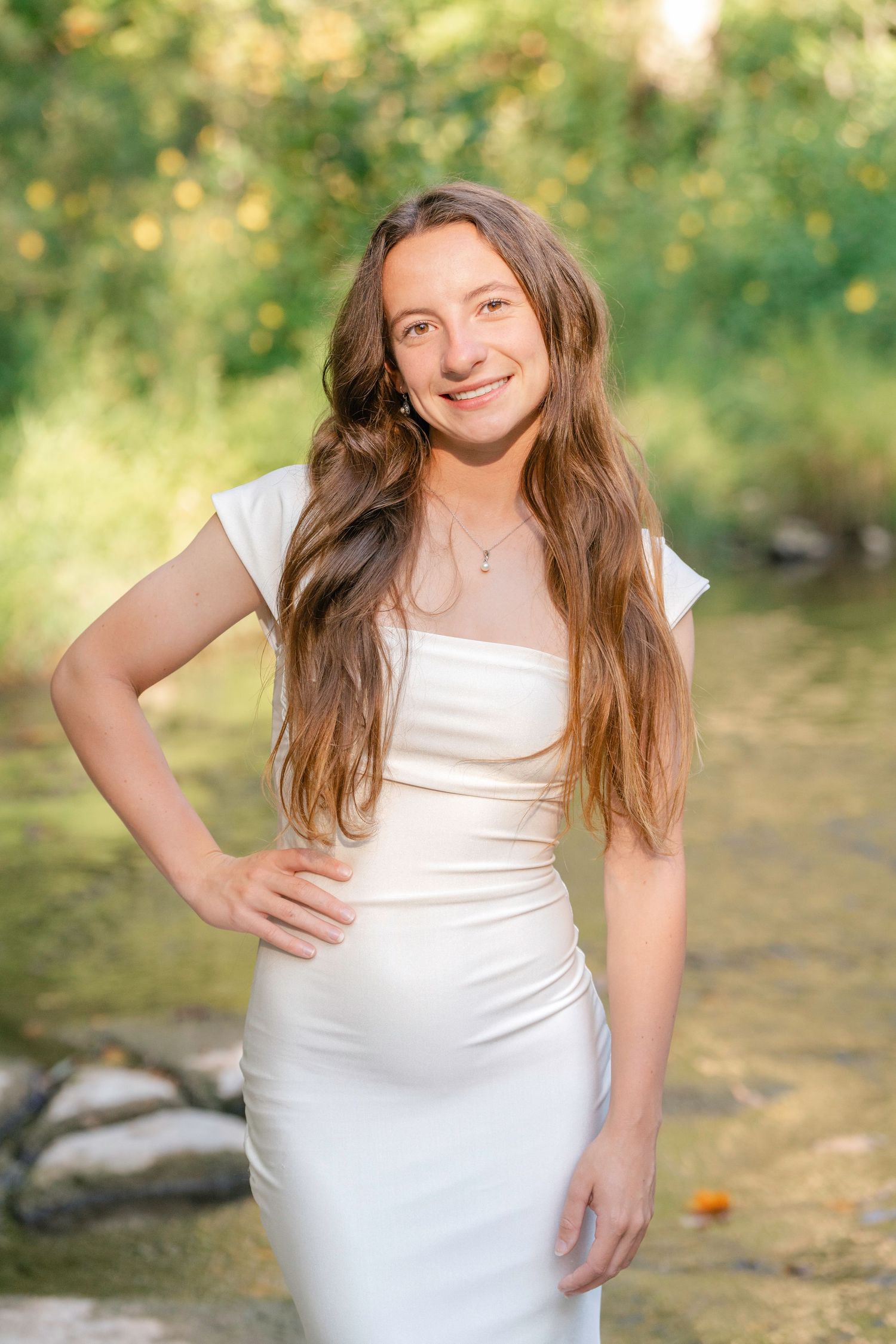 A person in a white dress poses outdoors by a stream with sunlit greenery in the background.