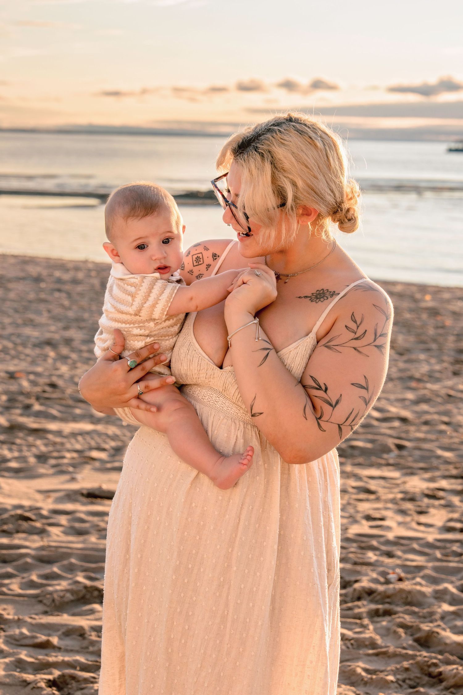 Person in cream dress holds and kisses baby on sandy beach at sunset.