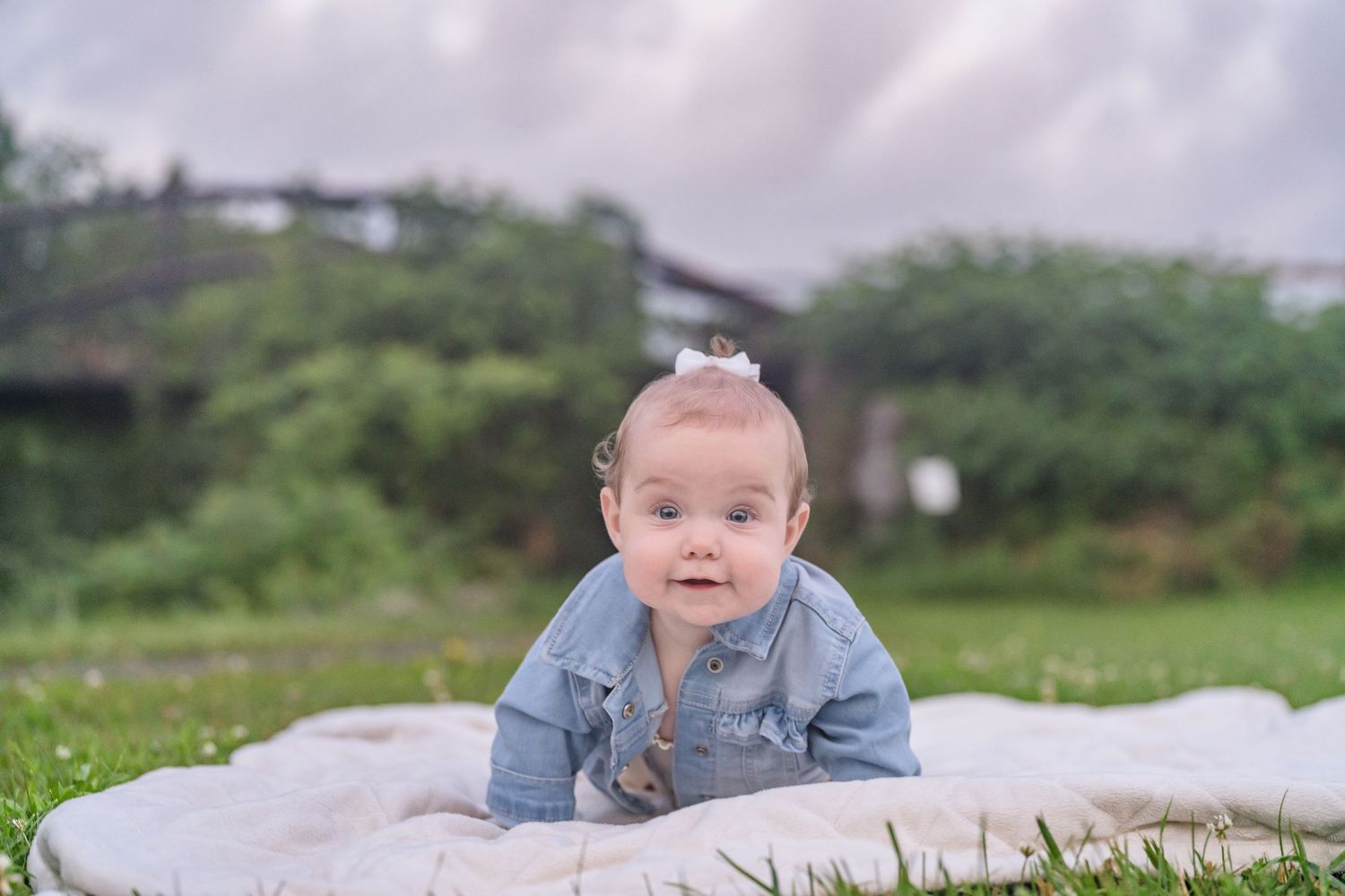Baby in denim jacket lies on white blanket in grassy outdoor setting.