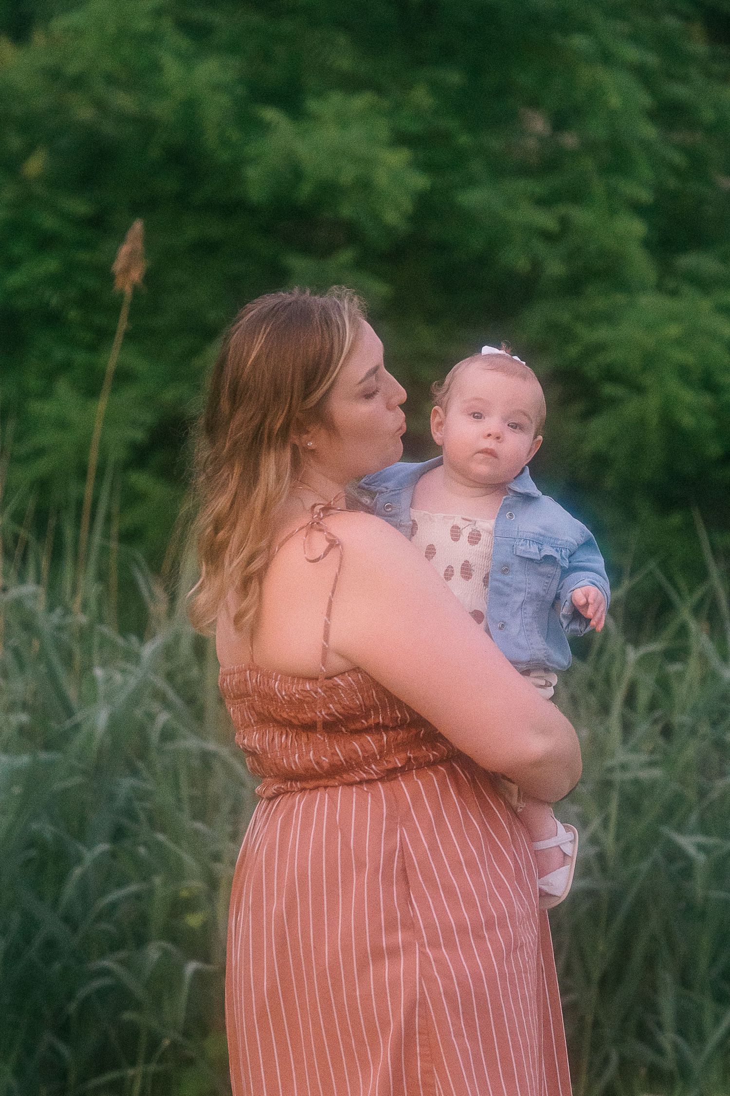 Mother in peach dress holds baby in denim jacket against green nature background.