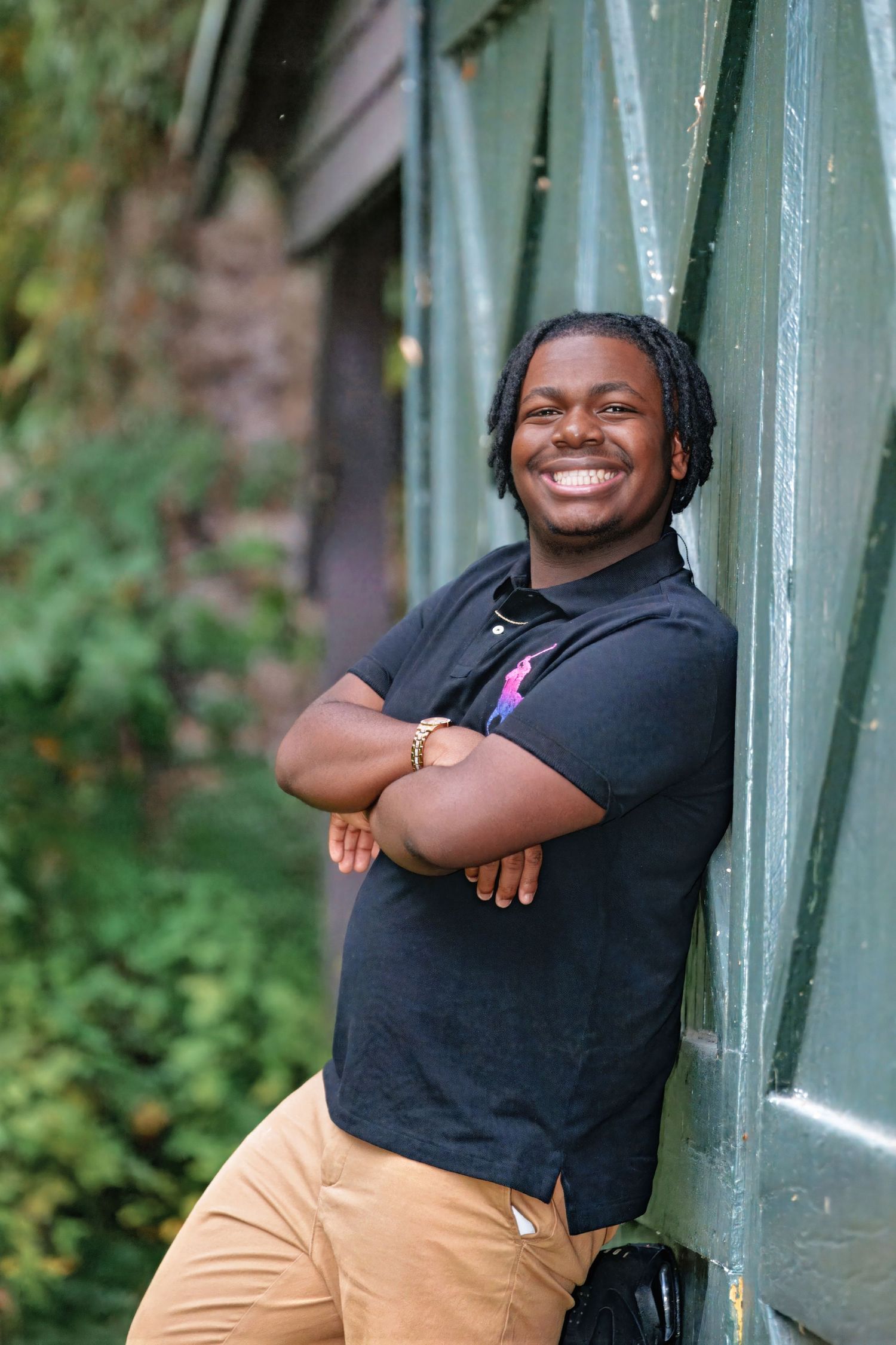 Person in black t-shirt and khaki pants poses joyfully against green metal structure in outdoor setting.