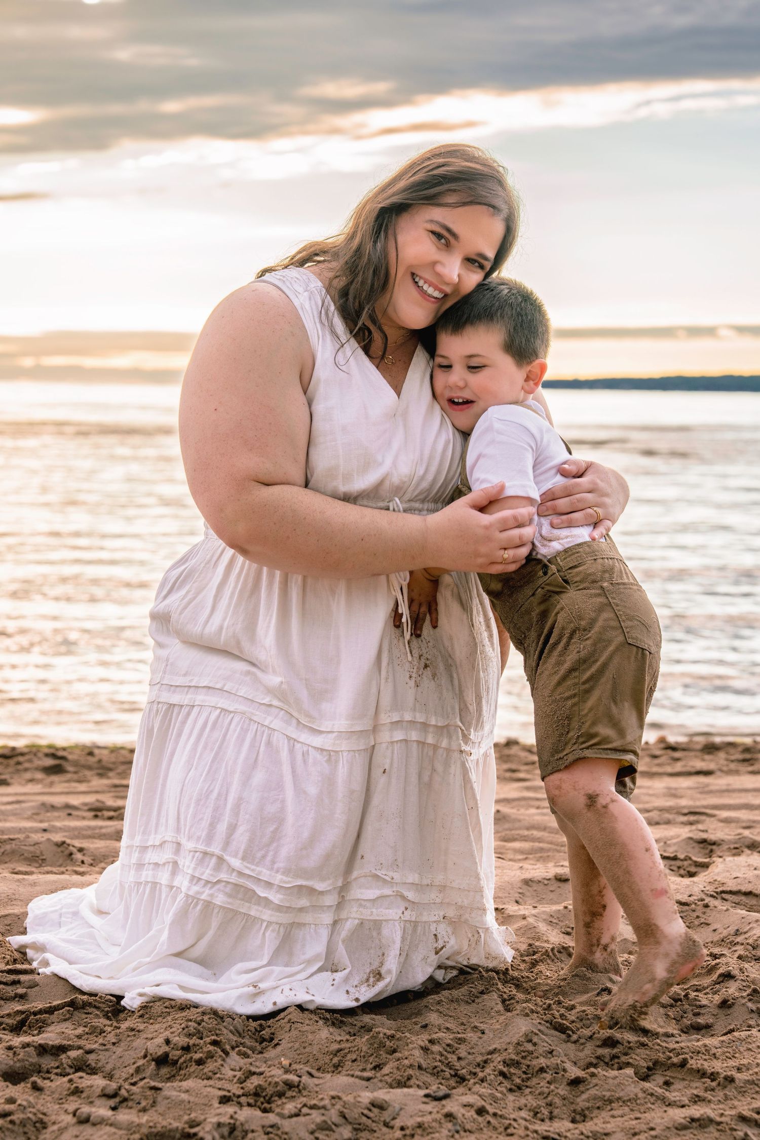 Mother and child in white dress and overalls share tender moment playing in sand at beach during sunset.