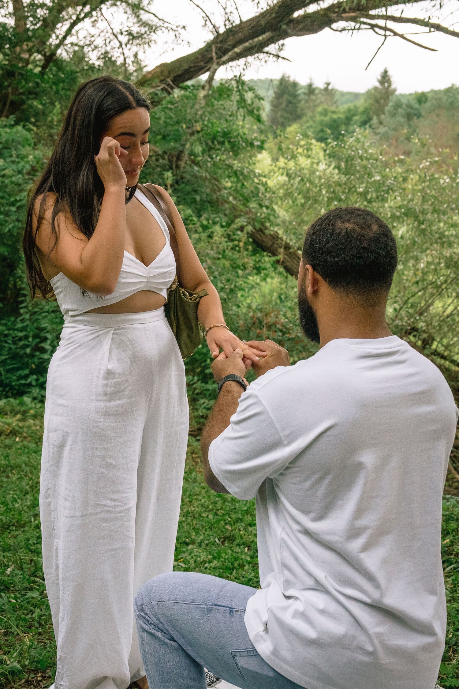 A couple shares a romantic moment in a lush green outdoor setting wearing white and casual attire.