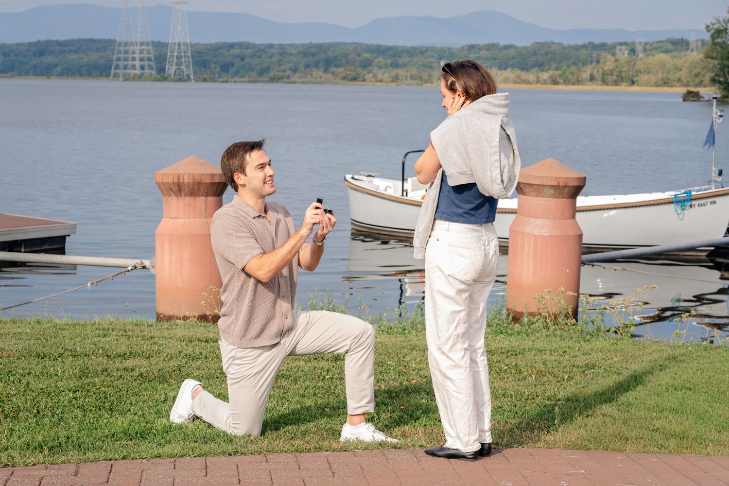 A romantic proposal scene unfolds on a lakeside dock with mountains and large terracotta planters in the background.