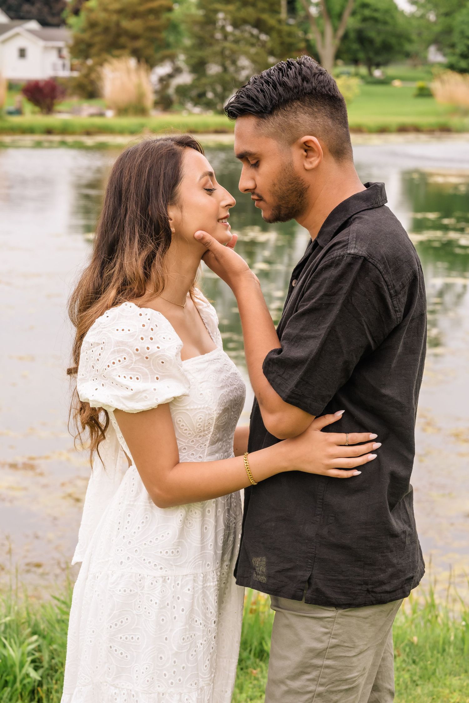 Couple shares intimate moment outdoors by lake with person in white eyelet dress and person in black shirt.