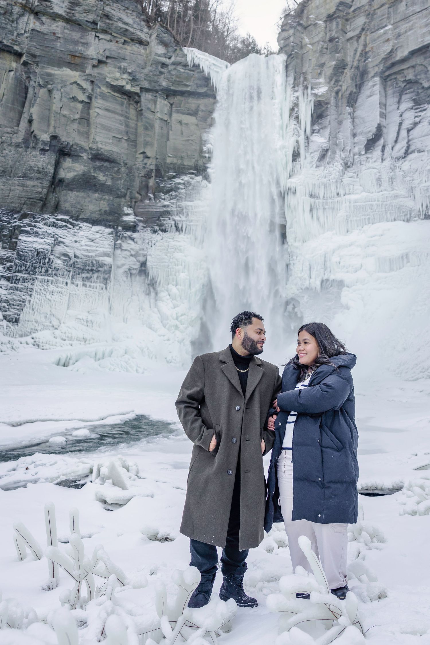 Two people share an intimate moment in front of a frozen waterfall surrounded by snow-covered cliffs.