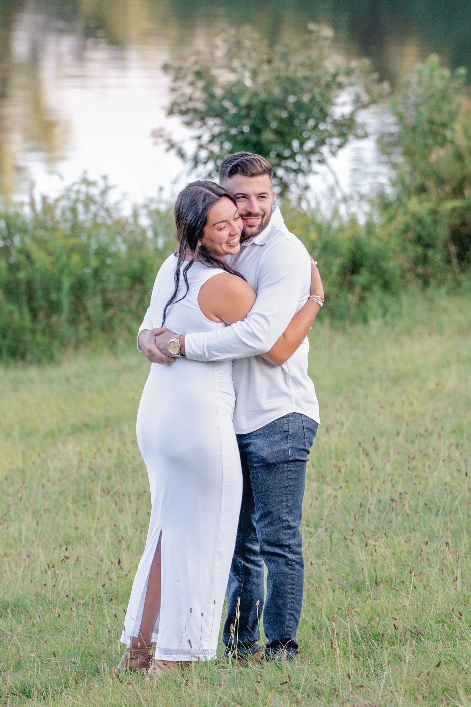 A couple shares a tender moment embracing on a grassy hillside near a lake during a summer day.