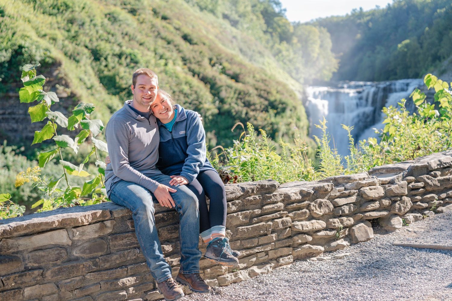 People sitting on stone wall overlooking scenic mountain waterfall and valley landscape in natural setting.