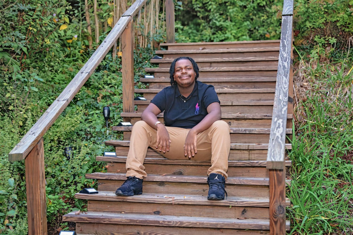 Person sits casually on wooden bench along outdoor trail surrounded by green foliage.