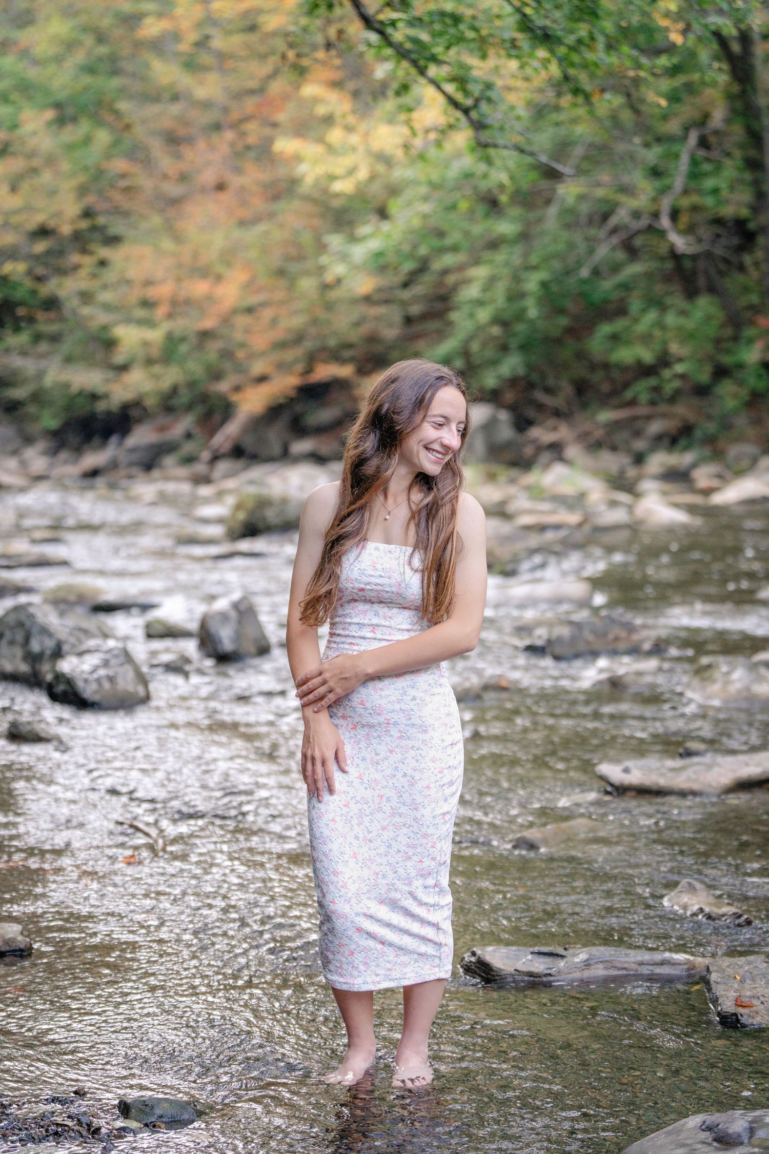 A person in a white dress stands along a rocky creek bed surrounded by autumn foliage.
