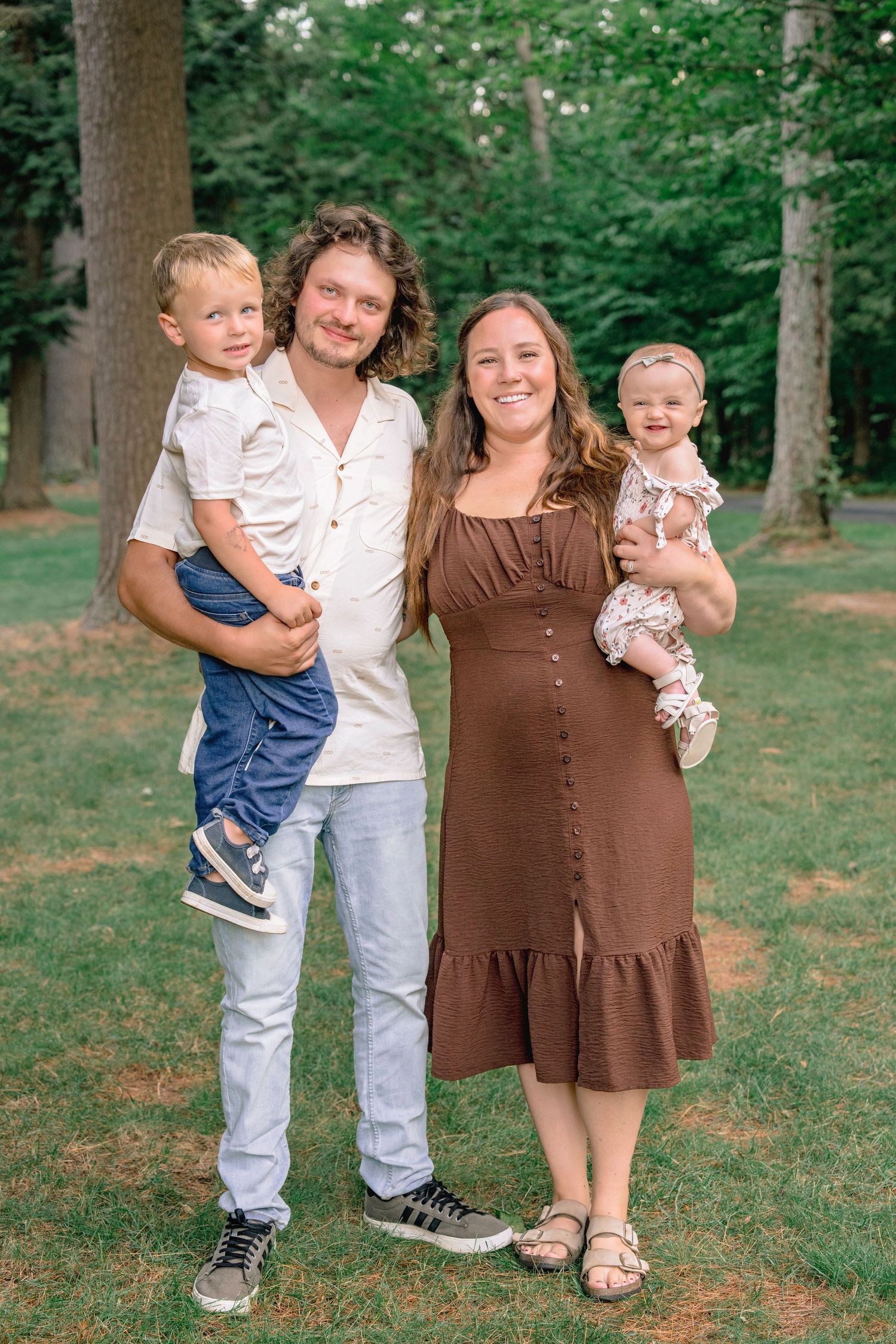 Family group poses together on grassy hill with trees in background wearing casual summer outfits.