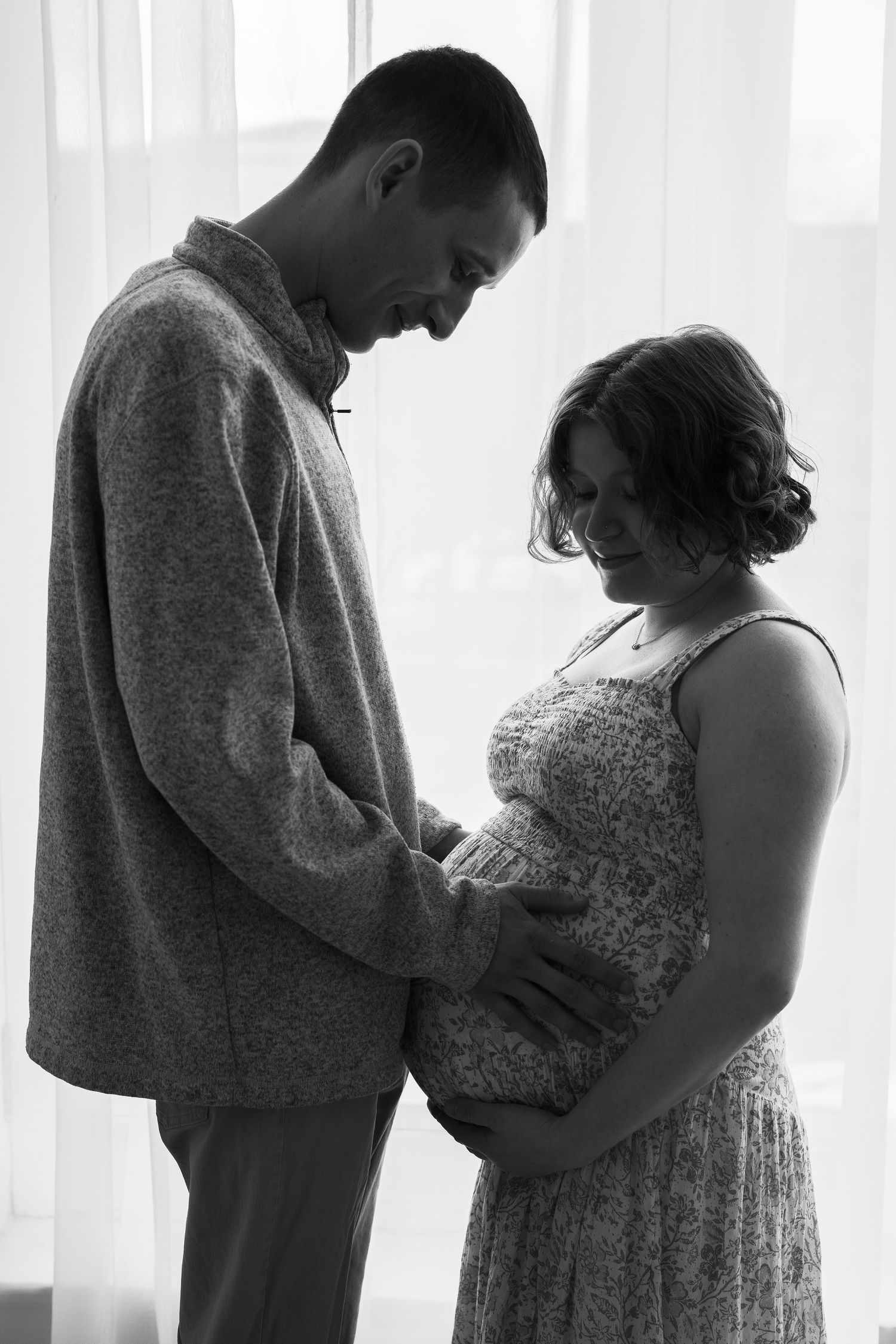 Black and white portrait of an intimate moment with couple cradling a pregnant belly in silhouette by window.