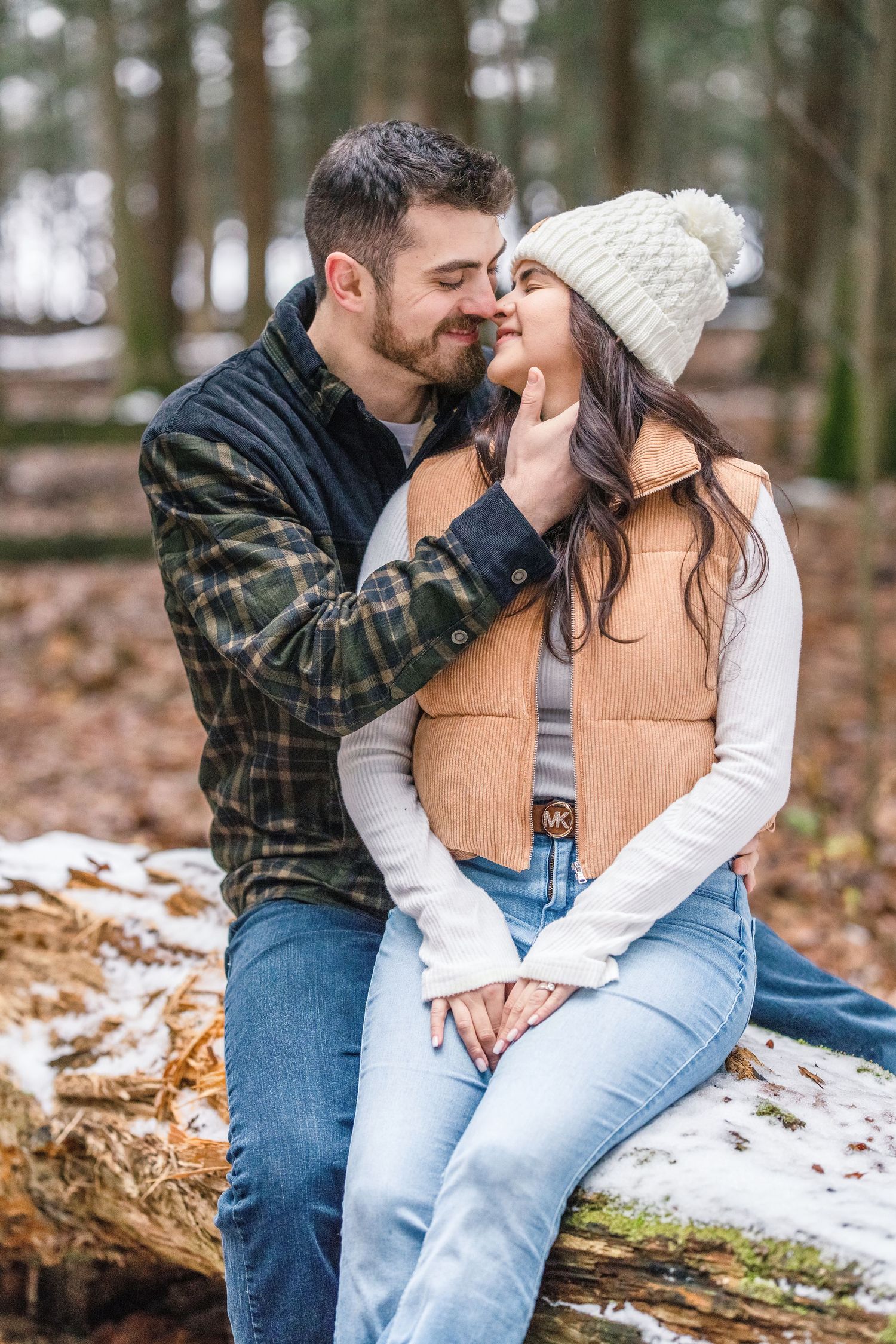 Couple sharing a romantic moment on a snowy log in winter woods, woman in beige vest and white knit hat.