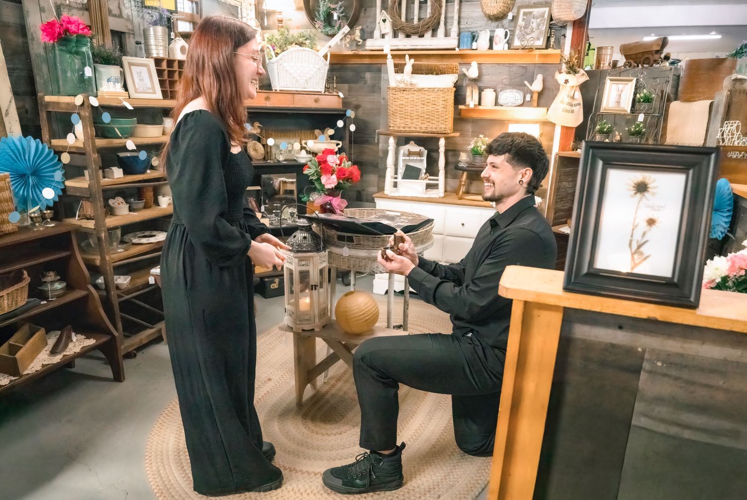 Man kneeling on one knee proposing to woman in black dress inside antique shop filled with decor.
