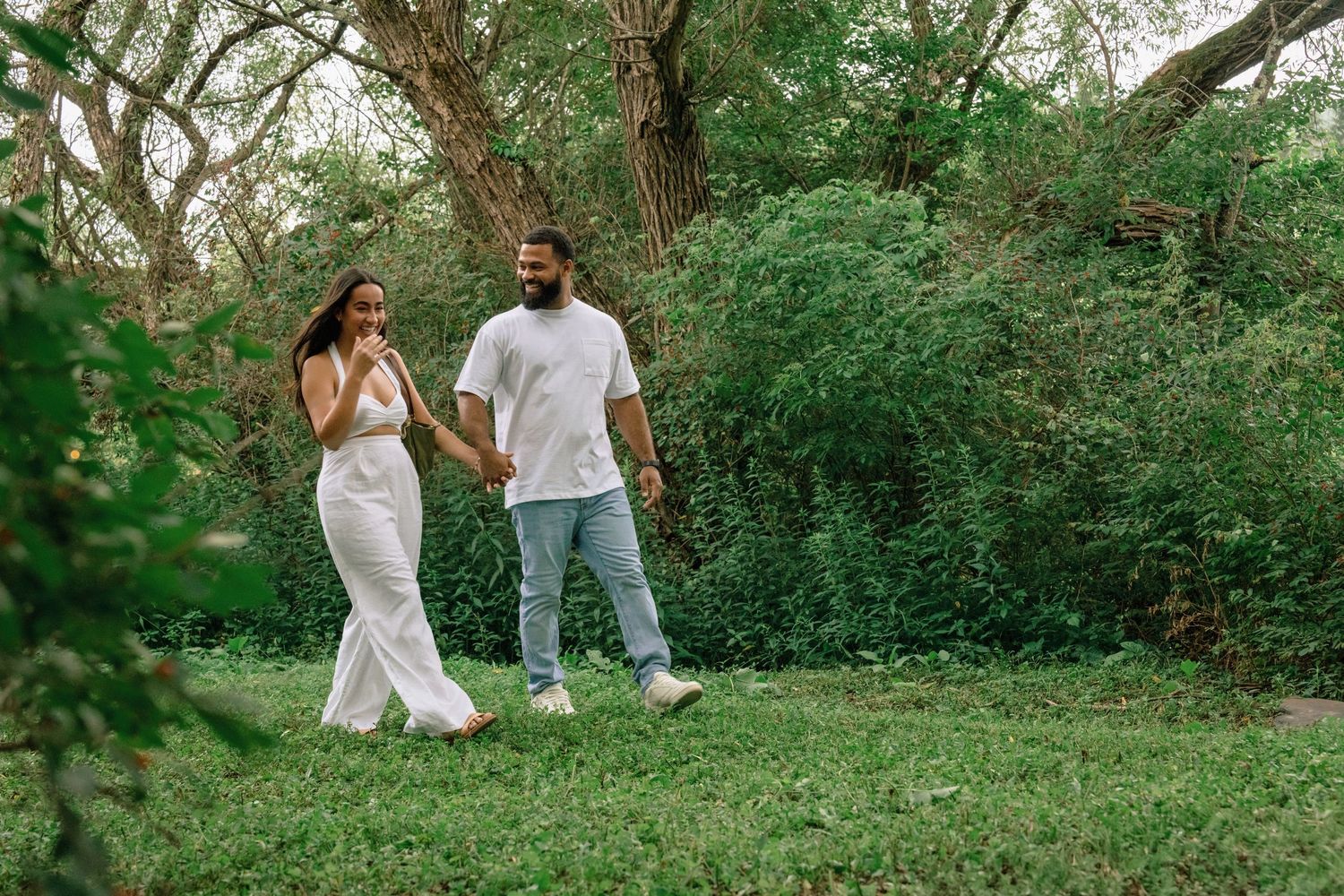 A couple walks hand in hand through a lush green park setting on a sunny day.