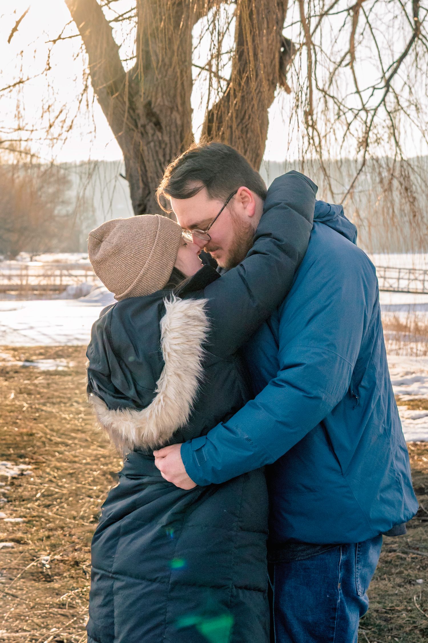 Couple embracing and kissing outdoors in winter near a bare tree in snow.