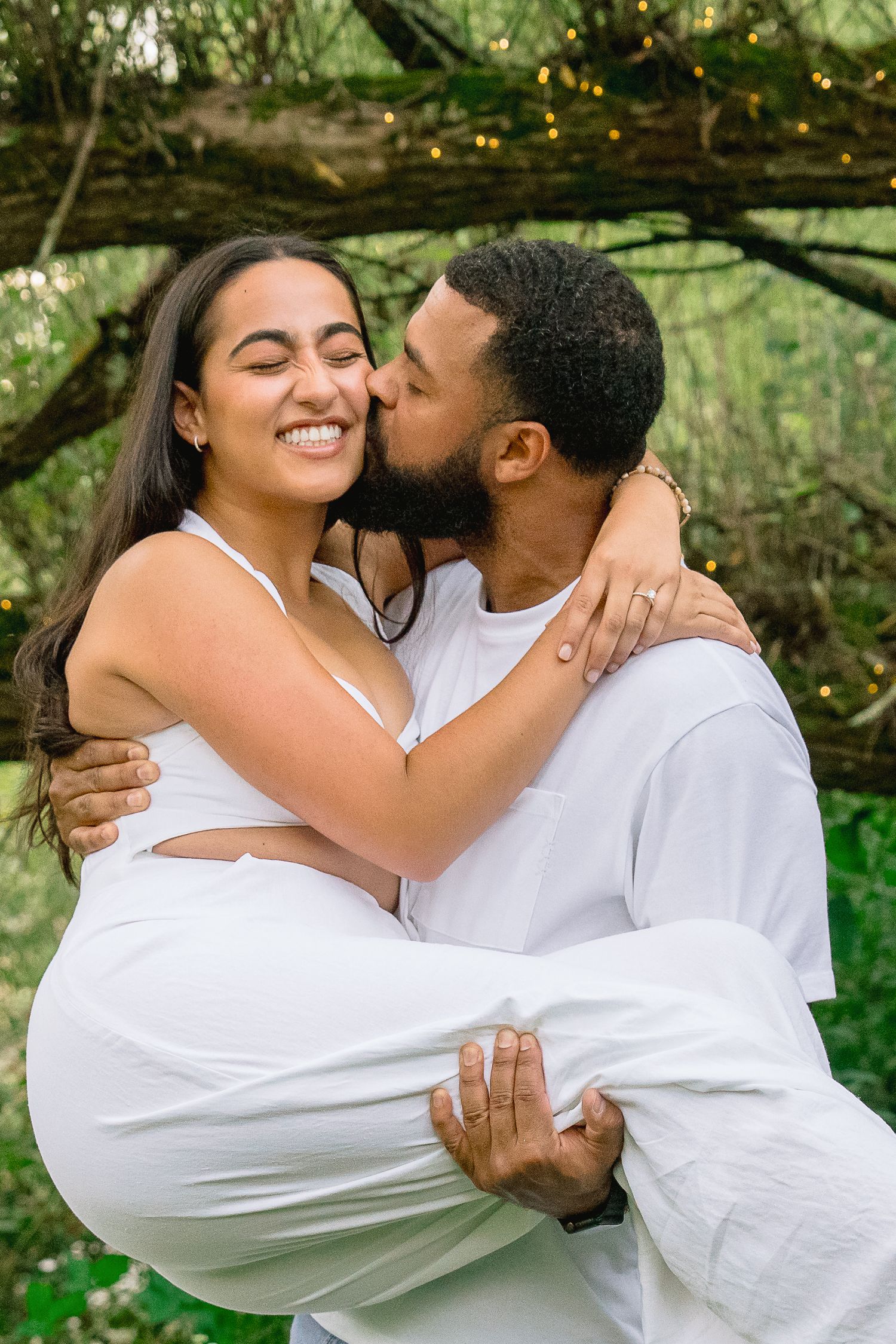 A romantic couple dressed in white embraces under a tree with twinkling lights in the background.