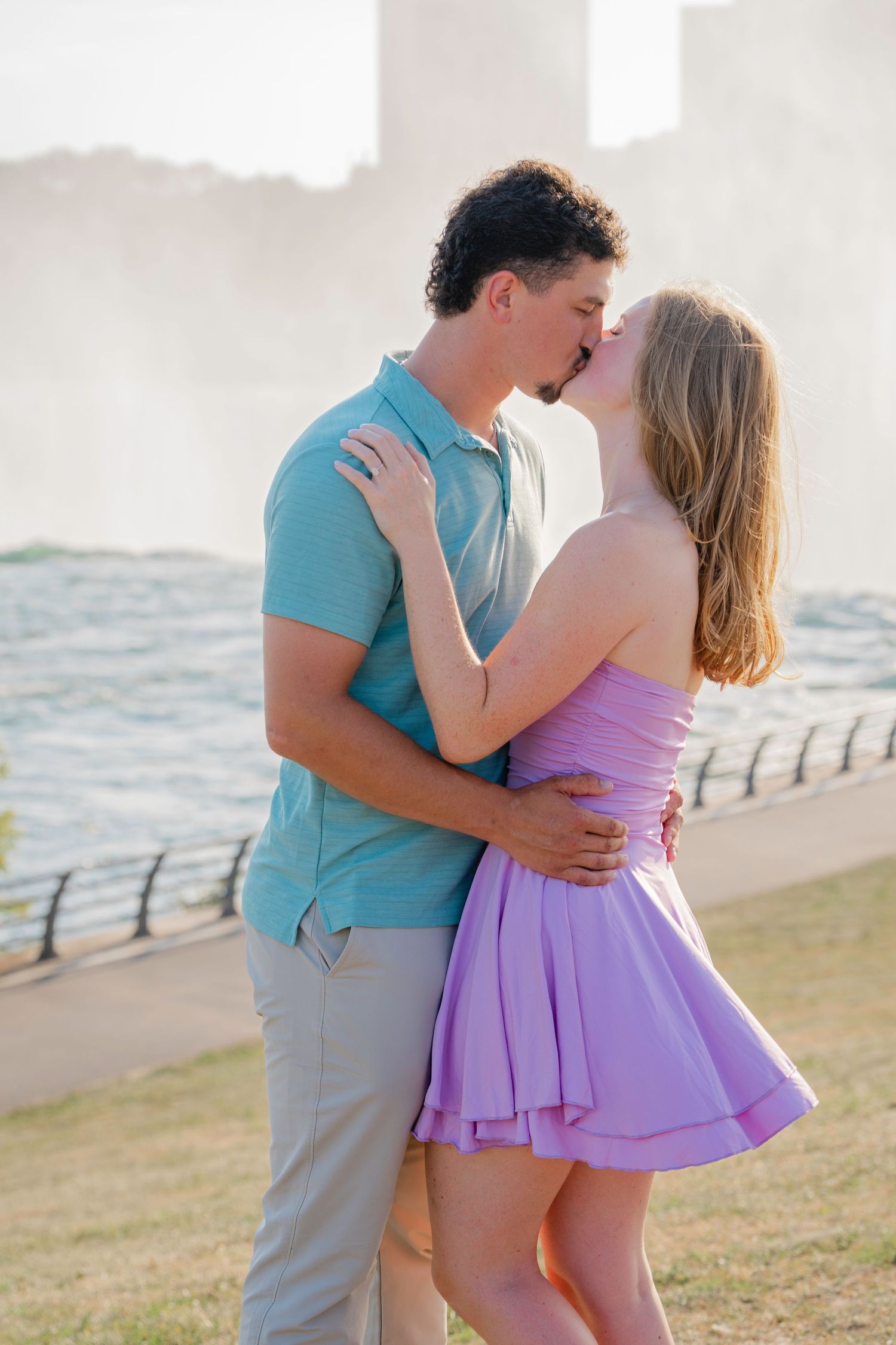 A romantic couple embraces by the waterfront, wearing coordinating turquoise and lavender outfits during sunset.