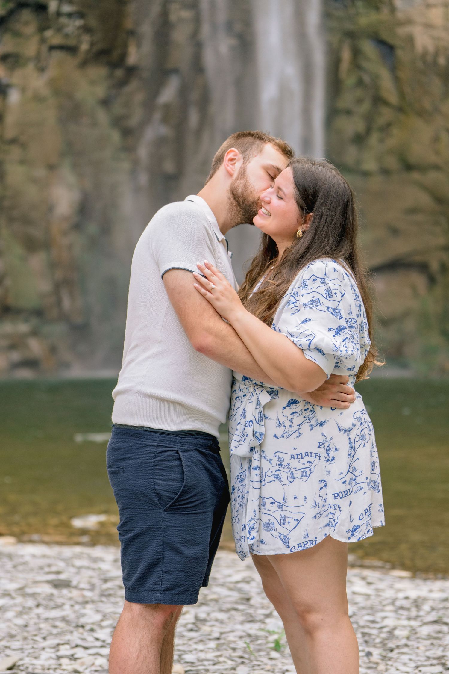 A romantic couple embraces near a waterfall with rocky cliffs and green water in the background.