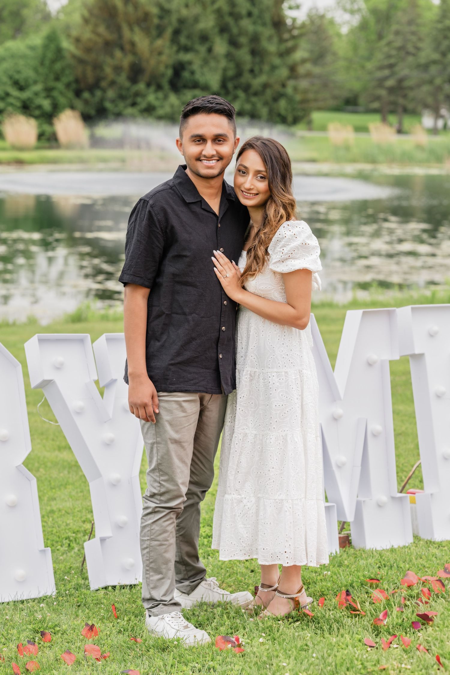 A couple poses together in front of white block letters on a grassy lawn near a pond.