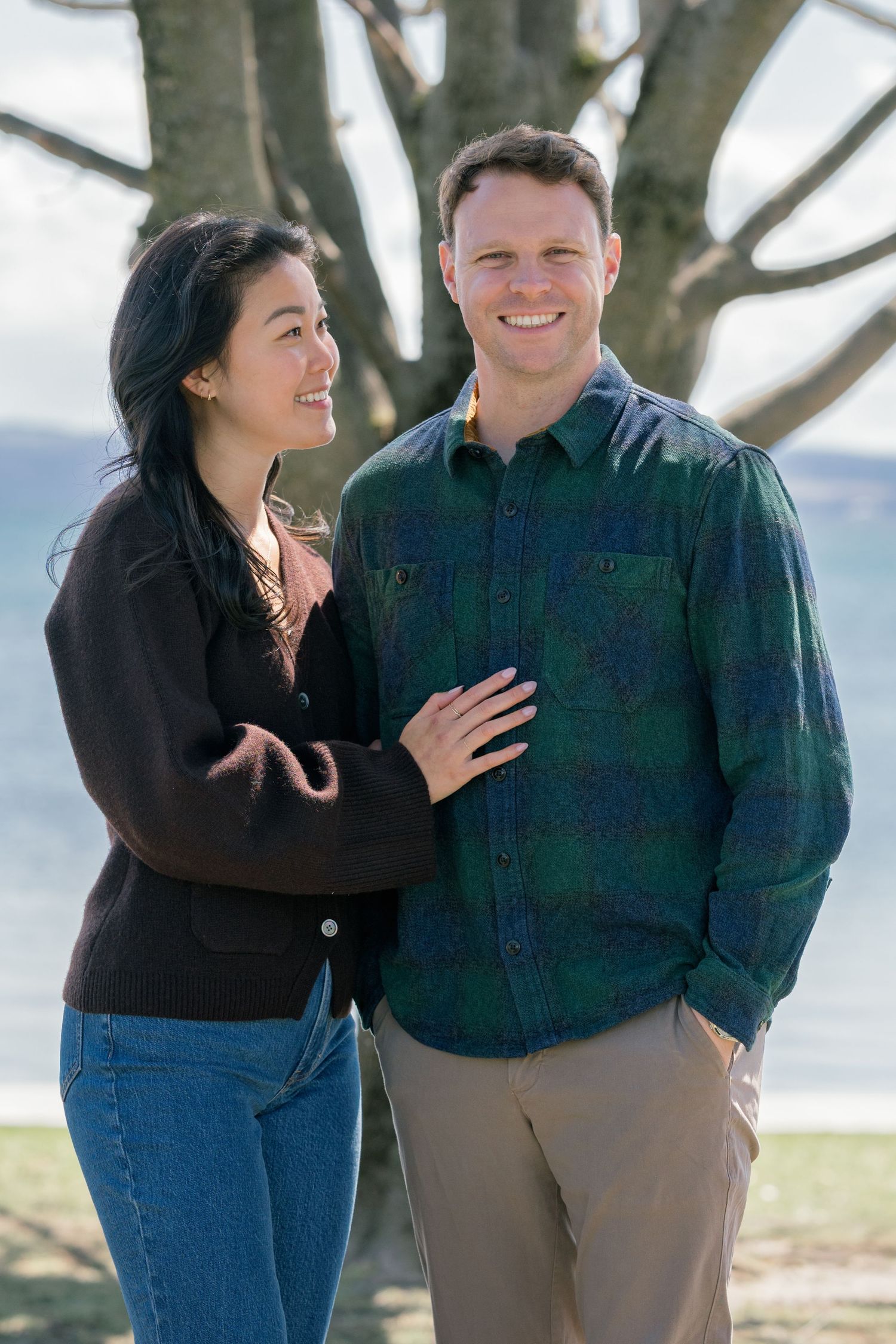 A couple stands together outdoors on a sunny day, with trees and water visible in the background.