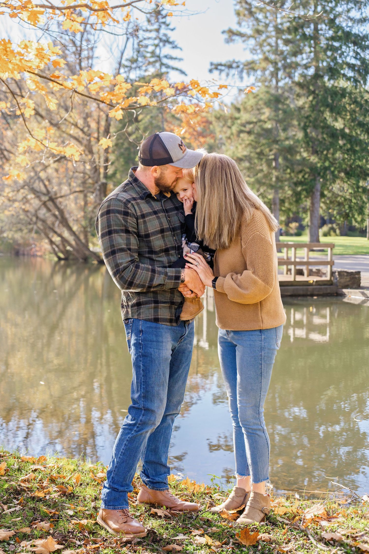 A couple shares a tender moment by a lake on an autumn day with fallen leaves and evergreen trees in the background.