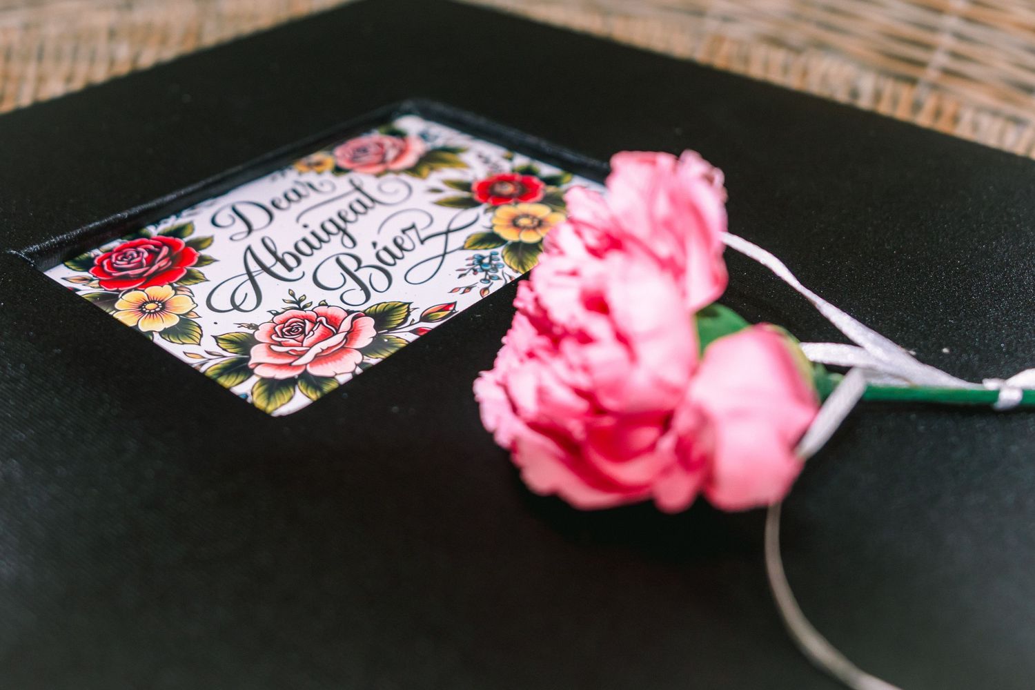 Floral place card reading Dear Abigail Baez on black surface with pink carnations and ribbon.