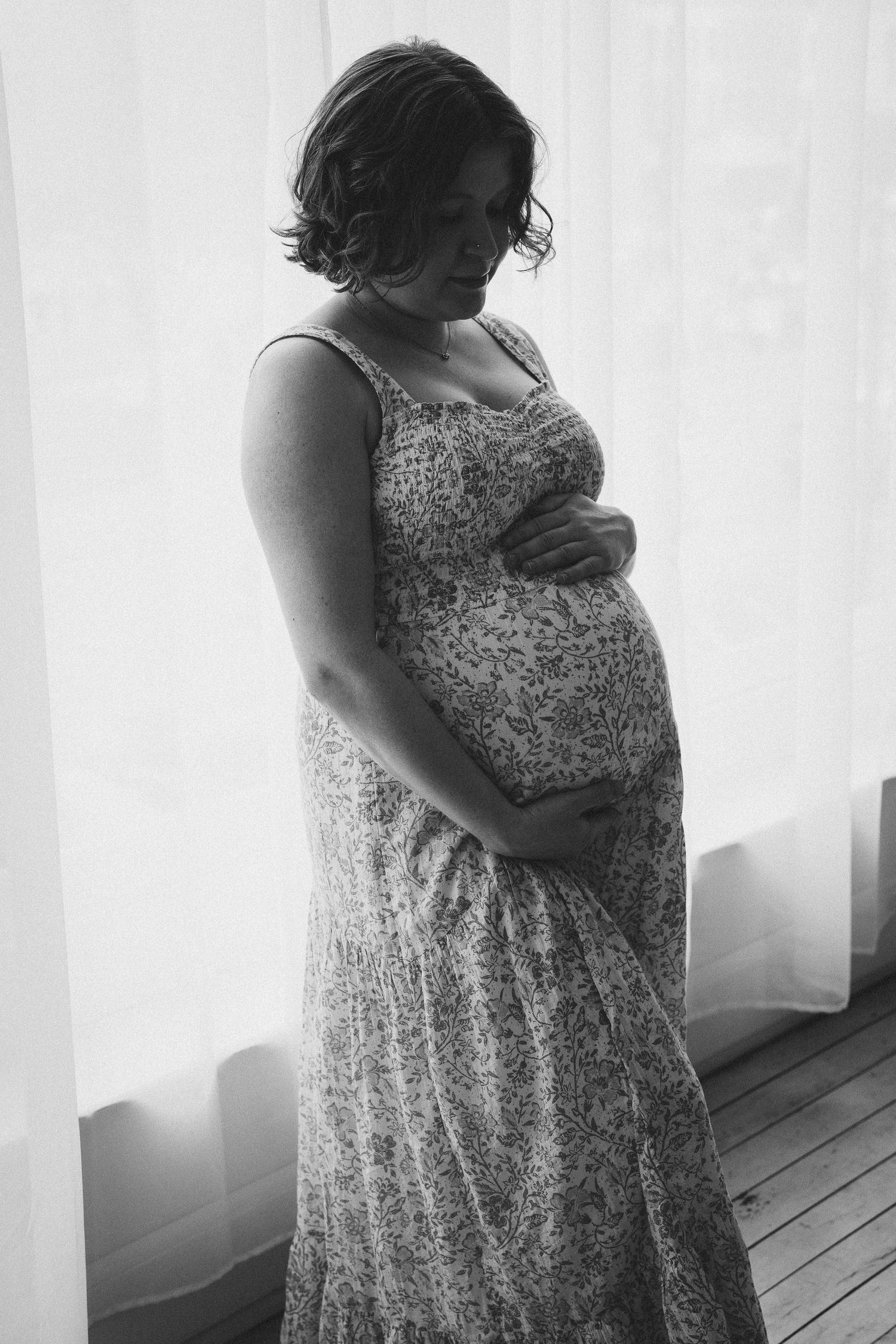 A side view of an expectant mother in a floral dress standing near a window, captured in black and white.