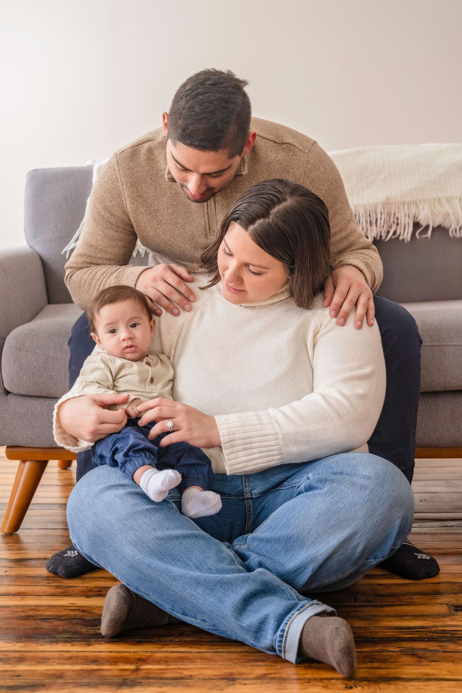 Family lounging together on hardwood floor with baby in cozy sweaters and jeans at home.