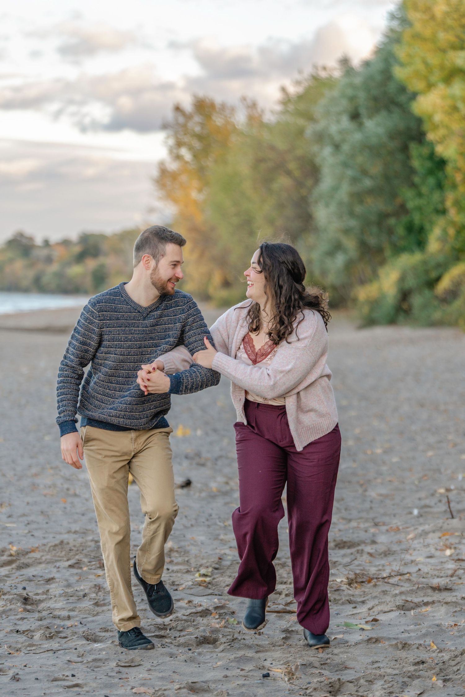 A playful couple runs and laughs together on a rocky beach during an autumn engagement session.