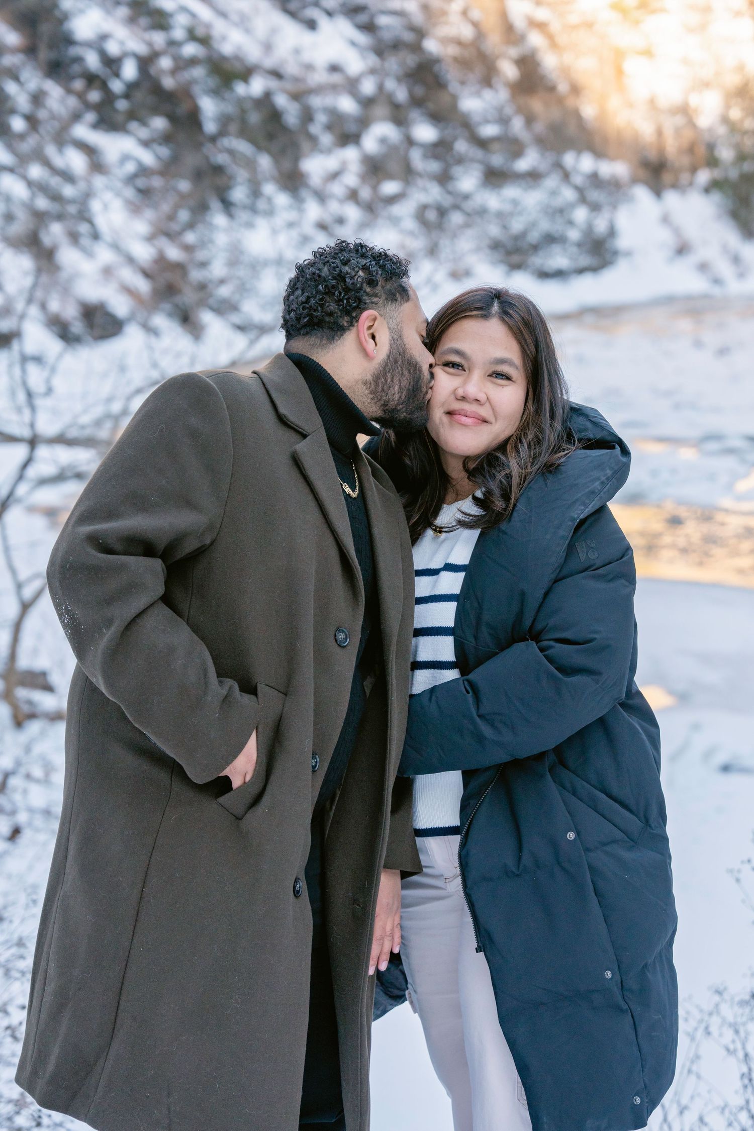 A couple shares tender moments in a snowy winter landscape during sunset while wearing warm coats.