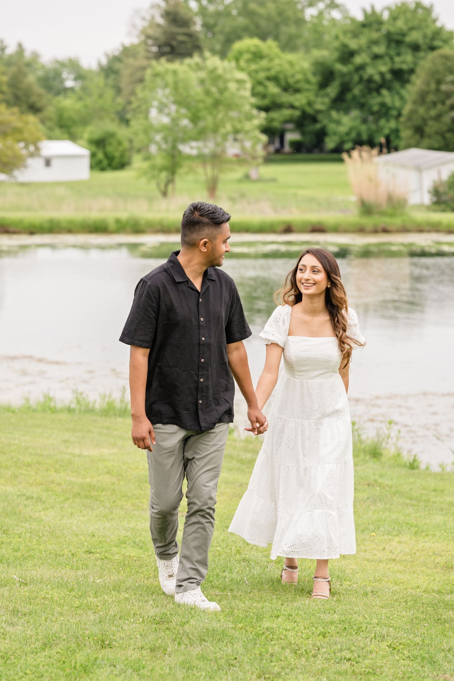 Couple takes a romantic stroll on grass near a lake wearing coordinated casual outfits.