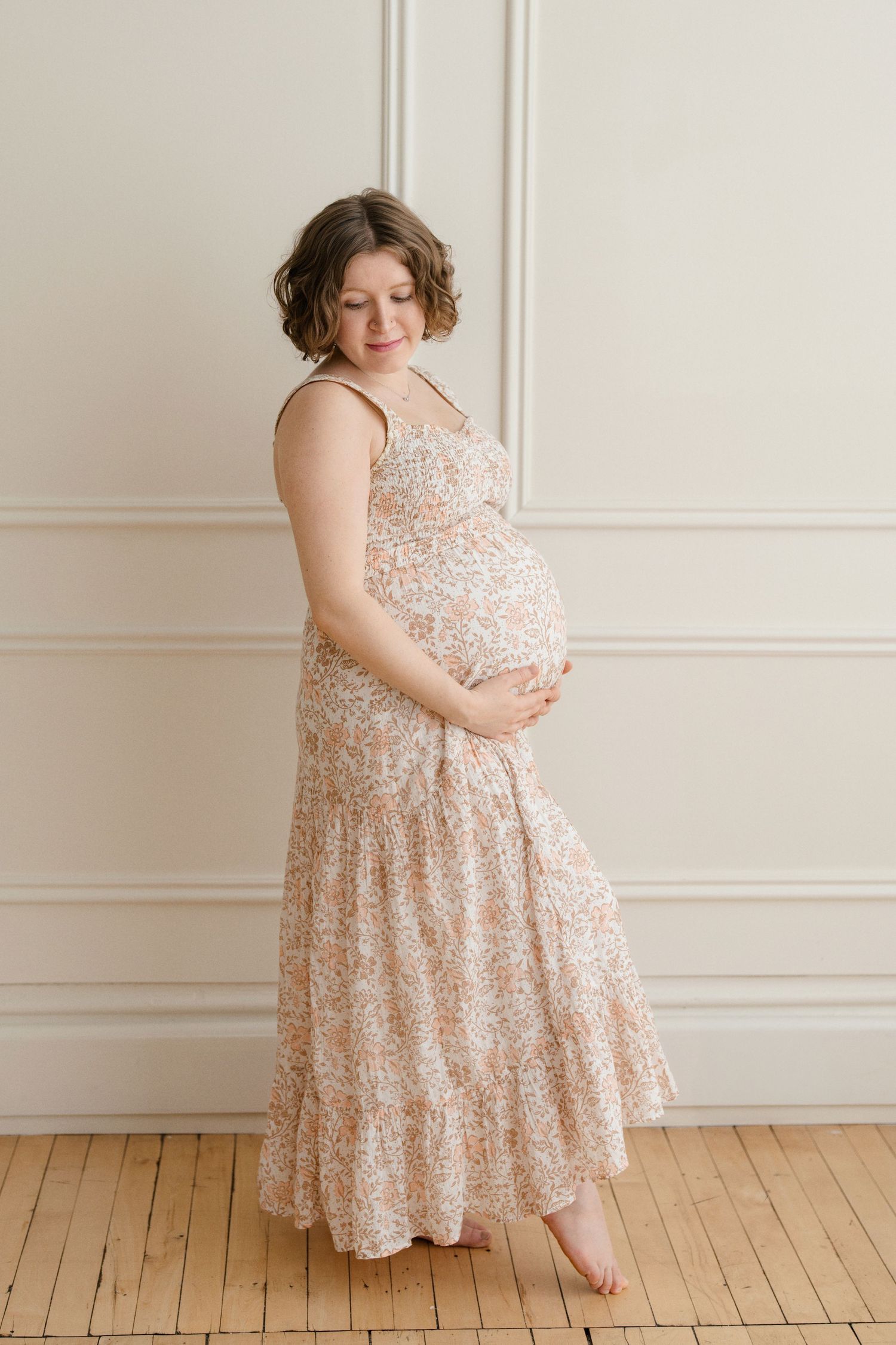 A pregnant woman in a floral maxi dress stands barefoot against a white paneled wall with hardwood floors.