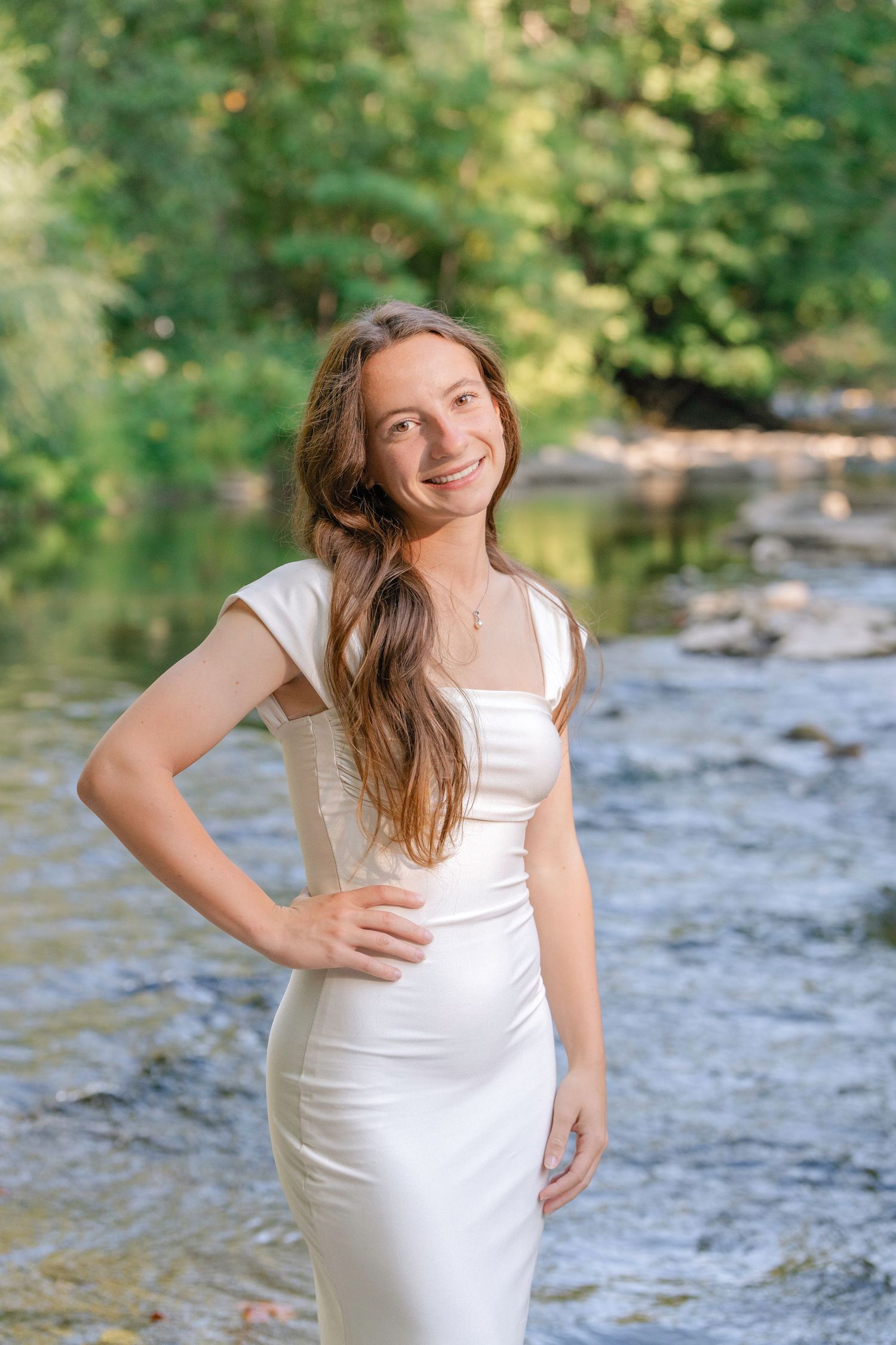 A person in a white dress poses gracefully by a flowing stream in a lush green forest setting.