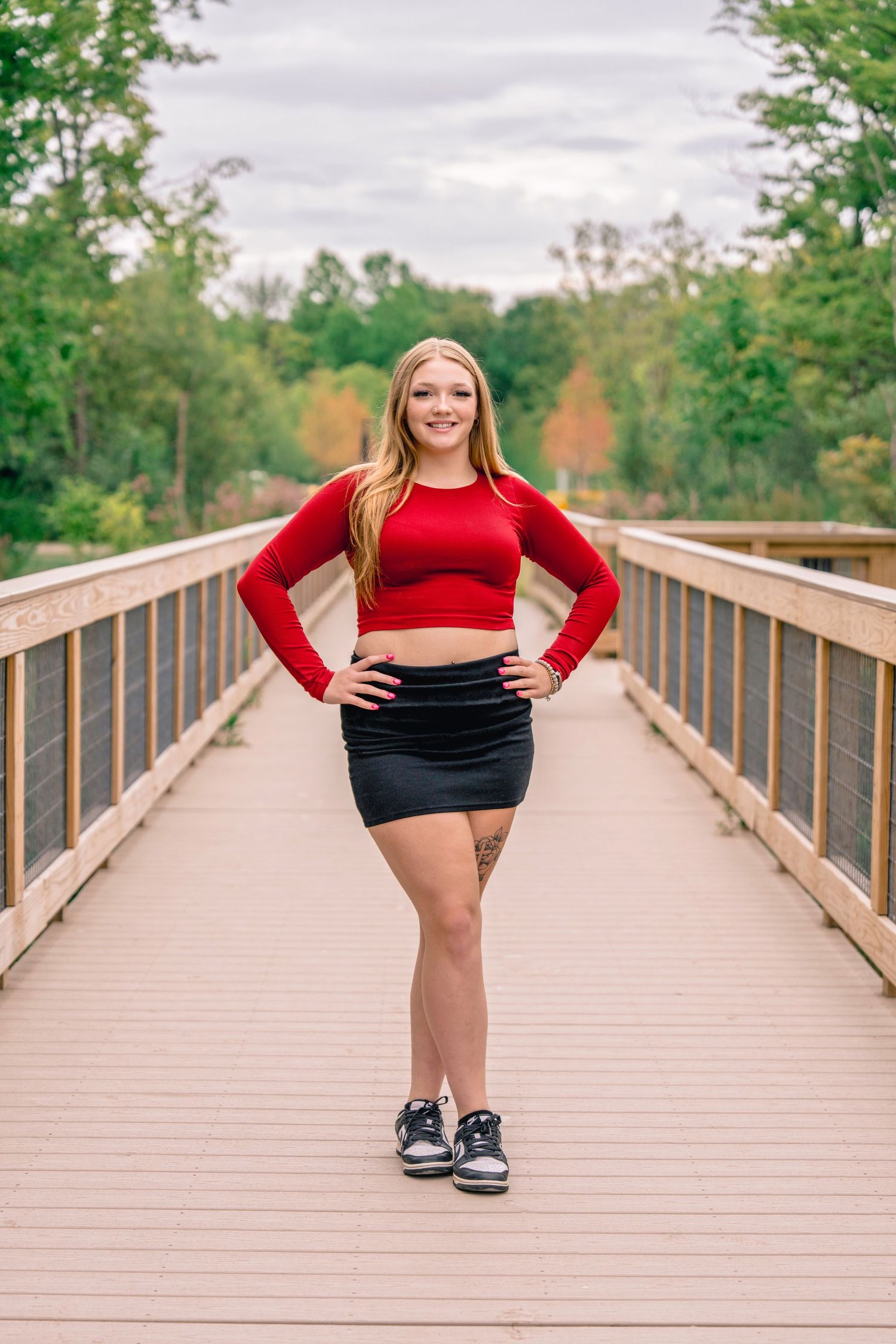 Person in red crop top and black skirt poses confidently on wooden boardwalk surrounded by trees.