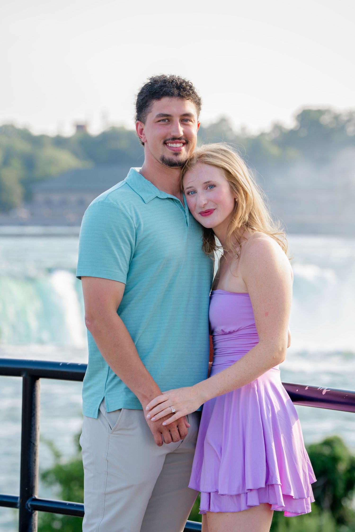 A couple embraces near a waterfall with a misty backdrop, wearing turquoise and purple outfits.