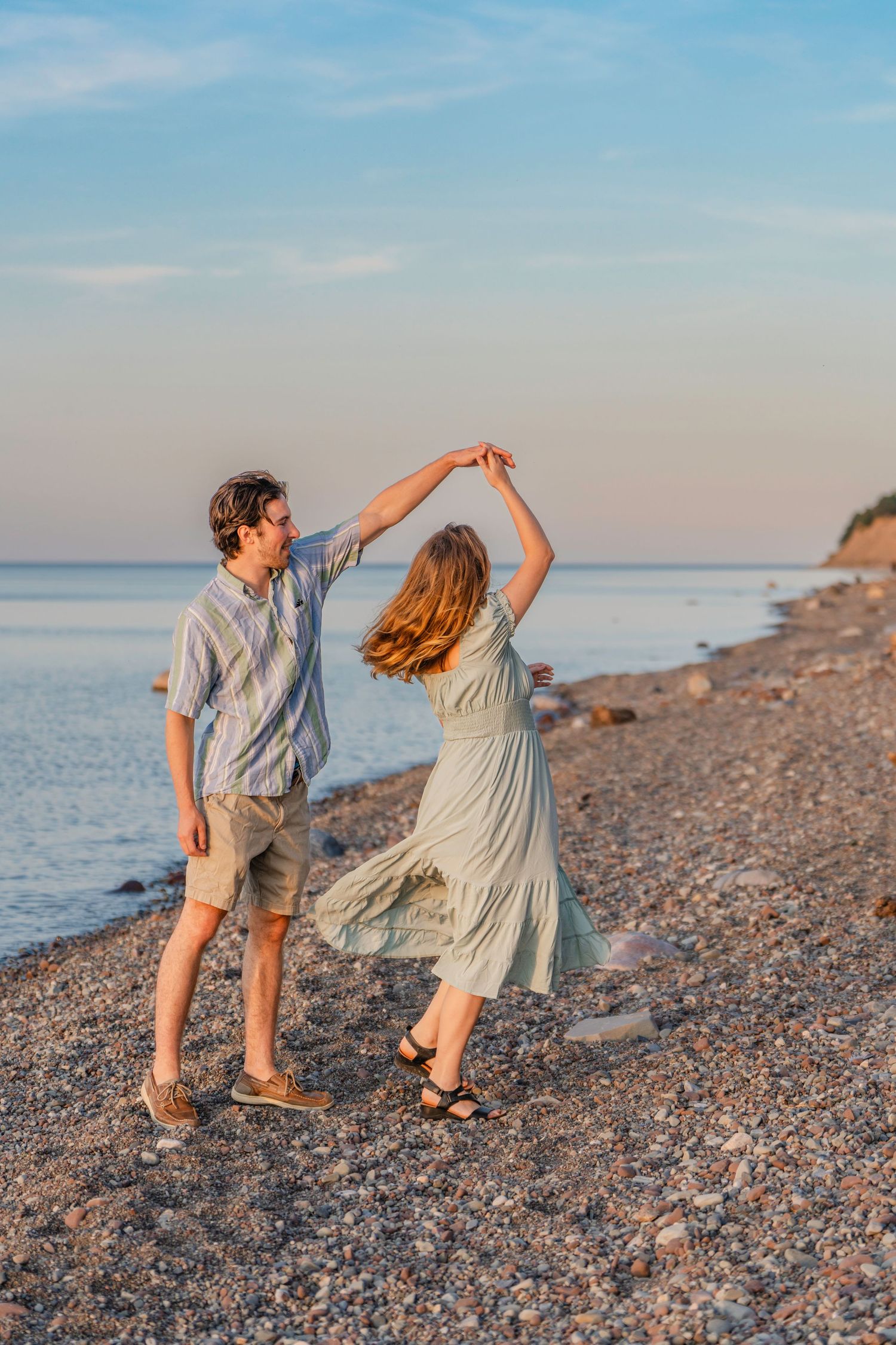 A couple dances together playfully on a rocky beach at sunset with the ocean in the background.
