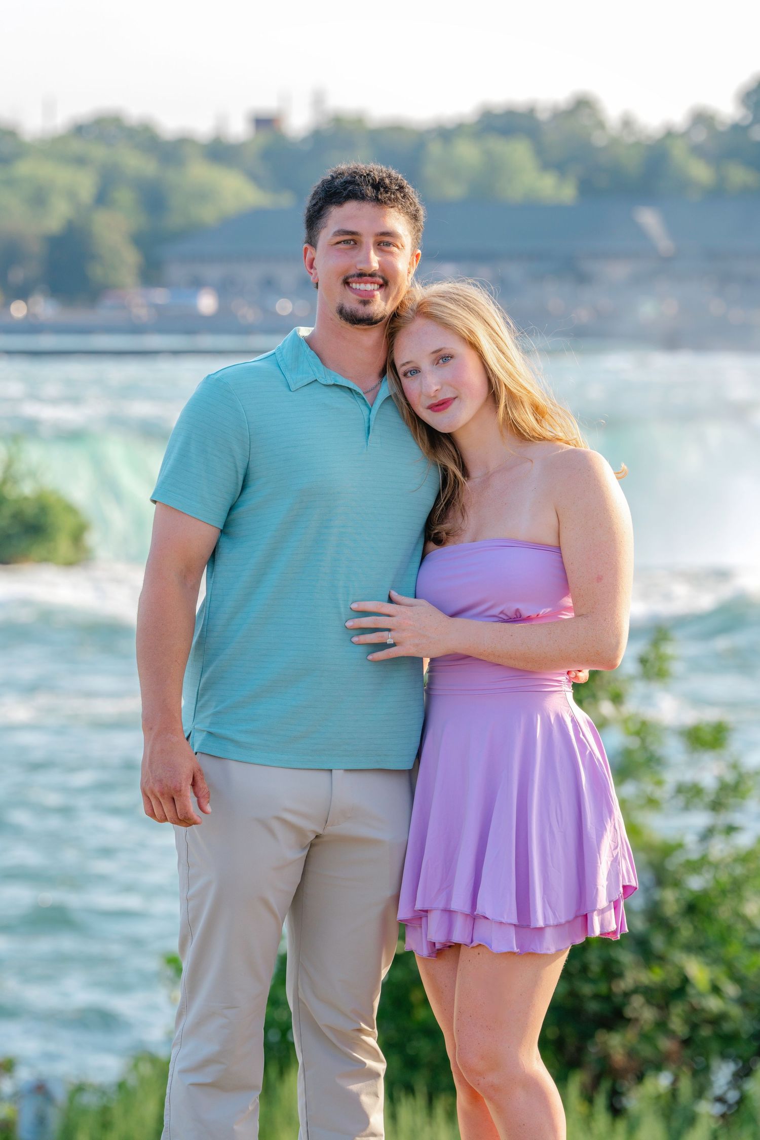 A couple poses together near Niagara Falls, wearing matching turquoise and purple outfits on a sunny day.