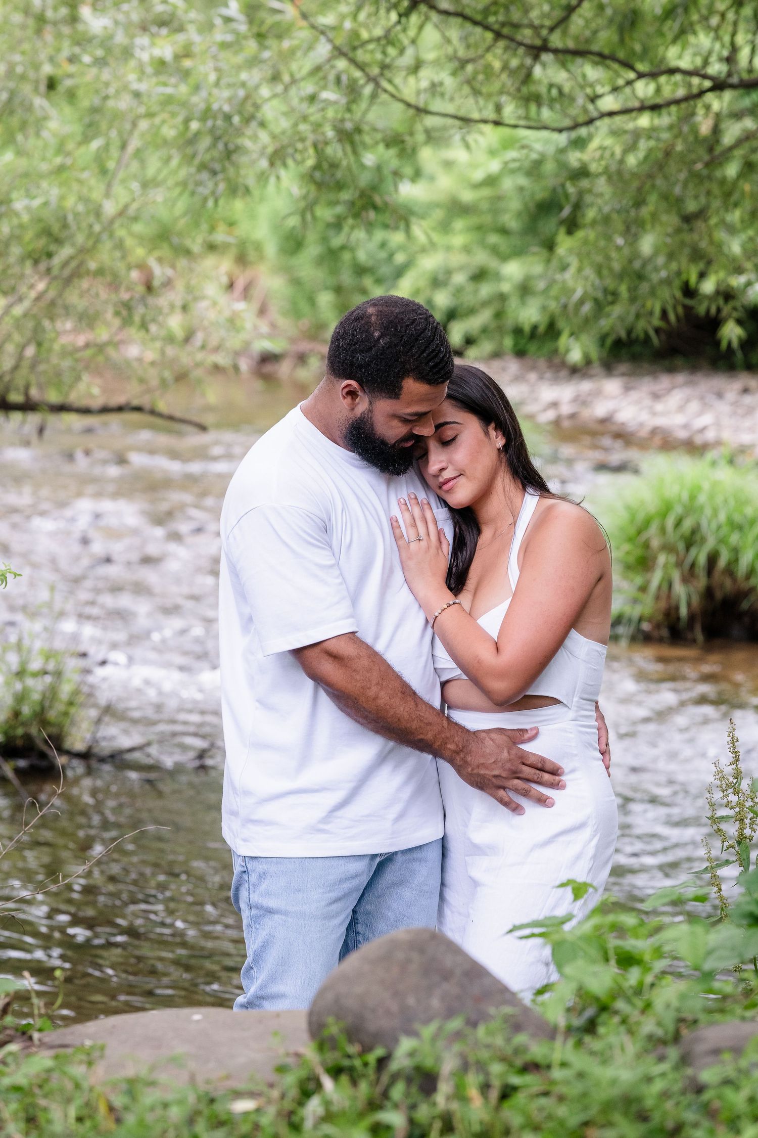 A romantic photoshoot by a creek with overhanging trees and natural scenery in the background.