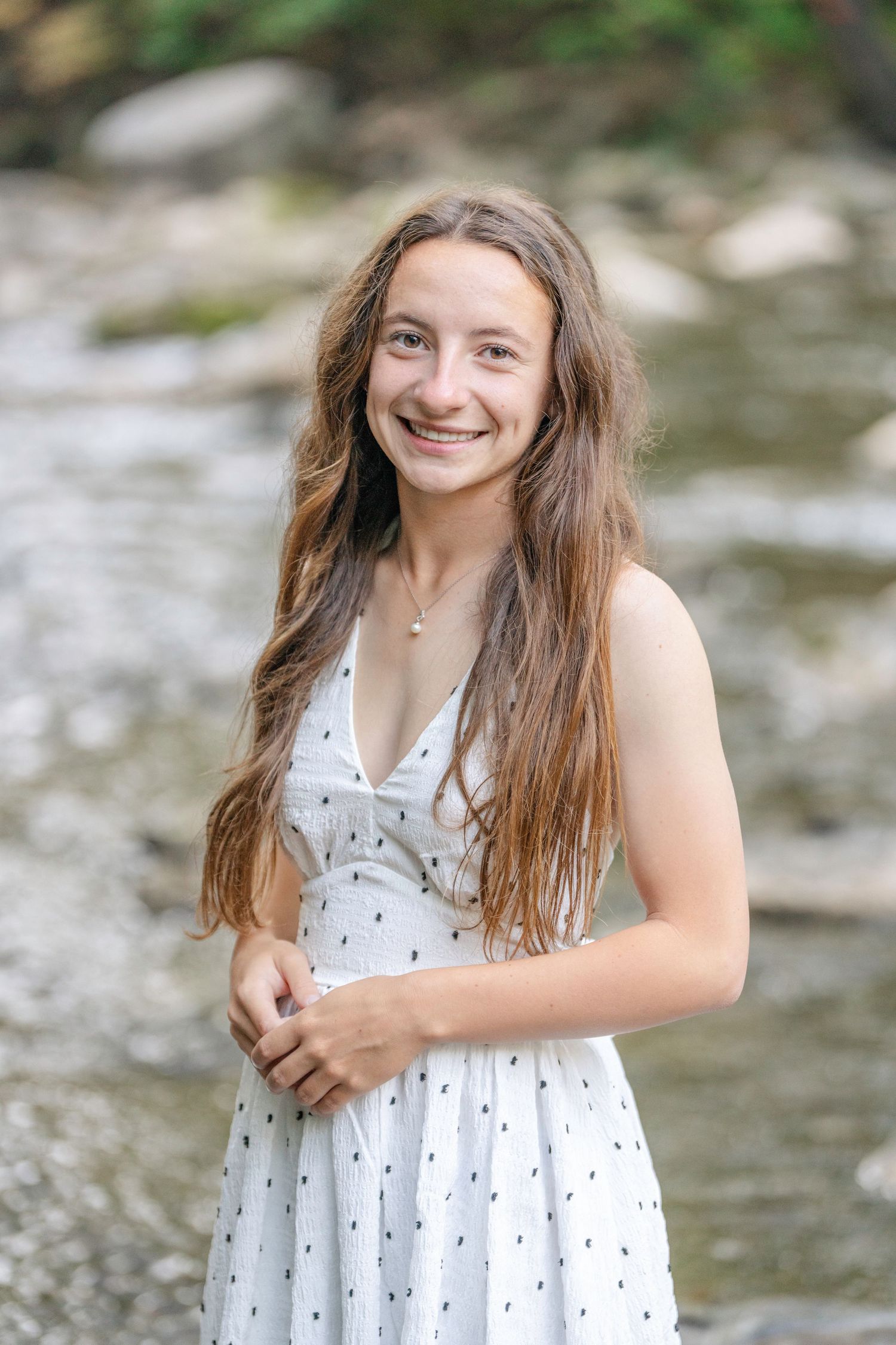 A young person in a white polka dot sundress smiles while standing near a rocky stream on a summer day.